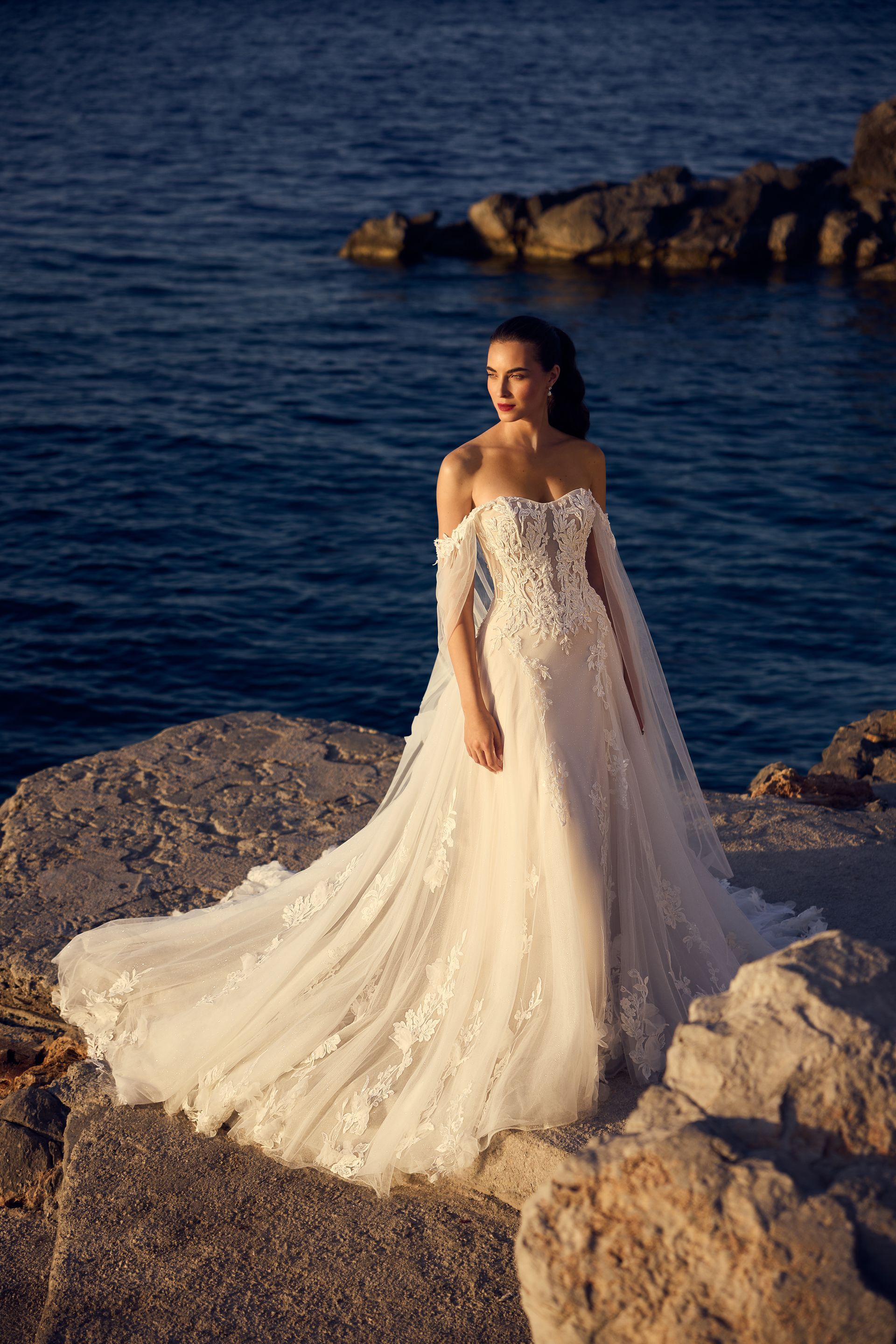 Woman in white wedding dress on rocky shore, ocean in the background.
