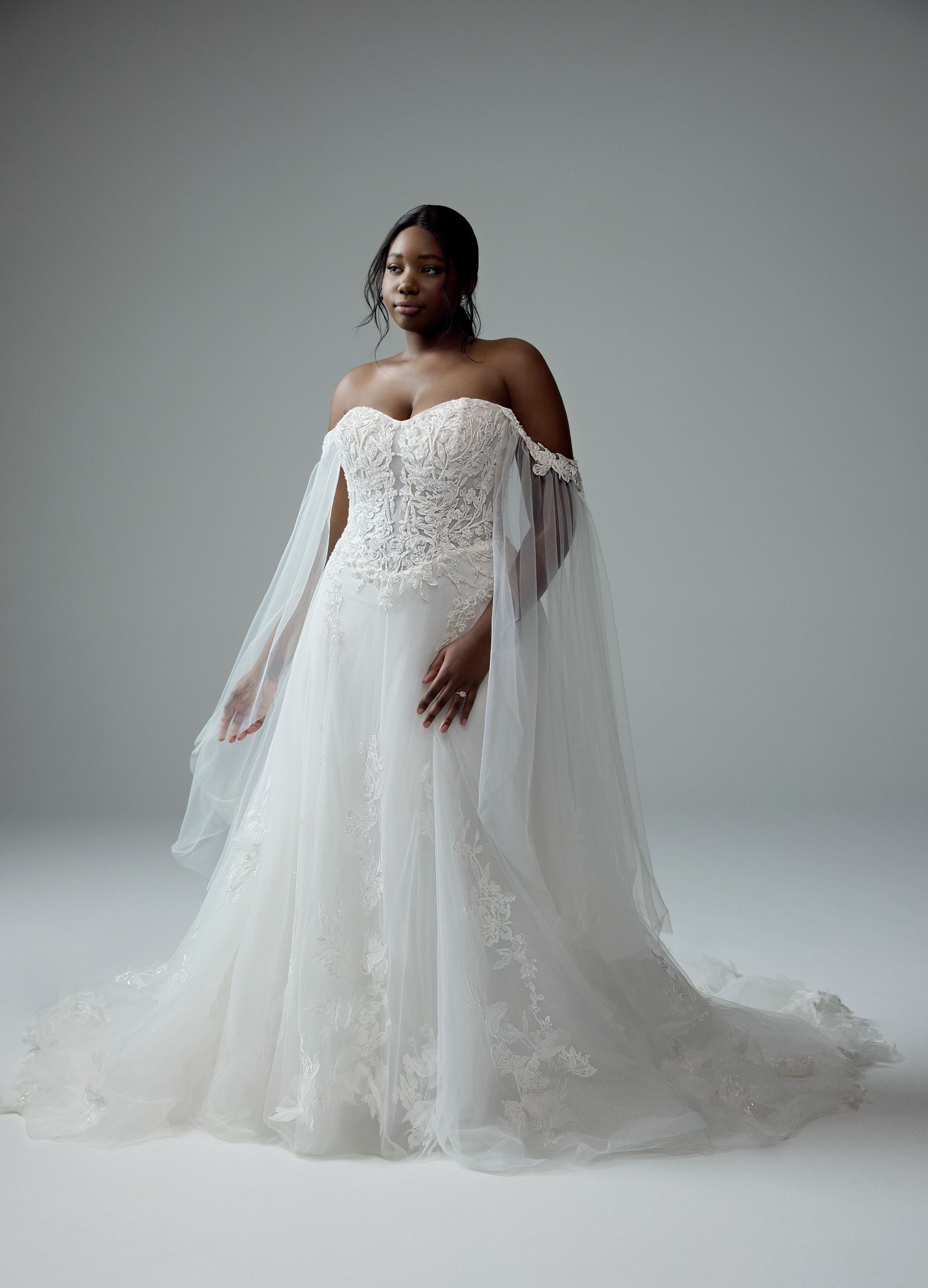 Woman in a white strapless wedding gown with flowing cape, posing in a studio.