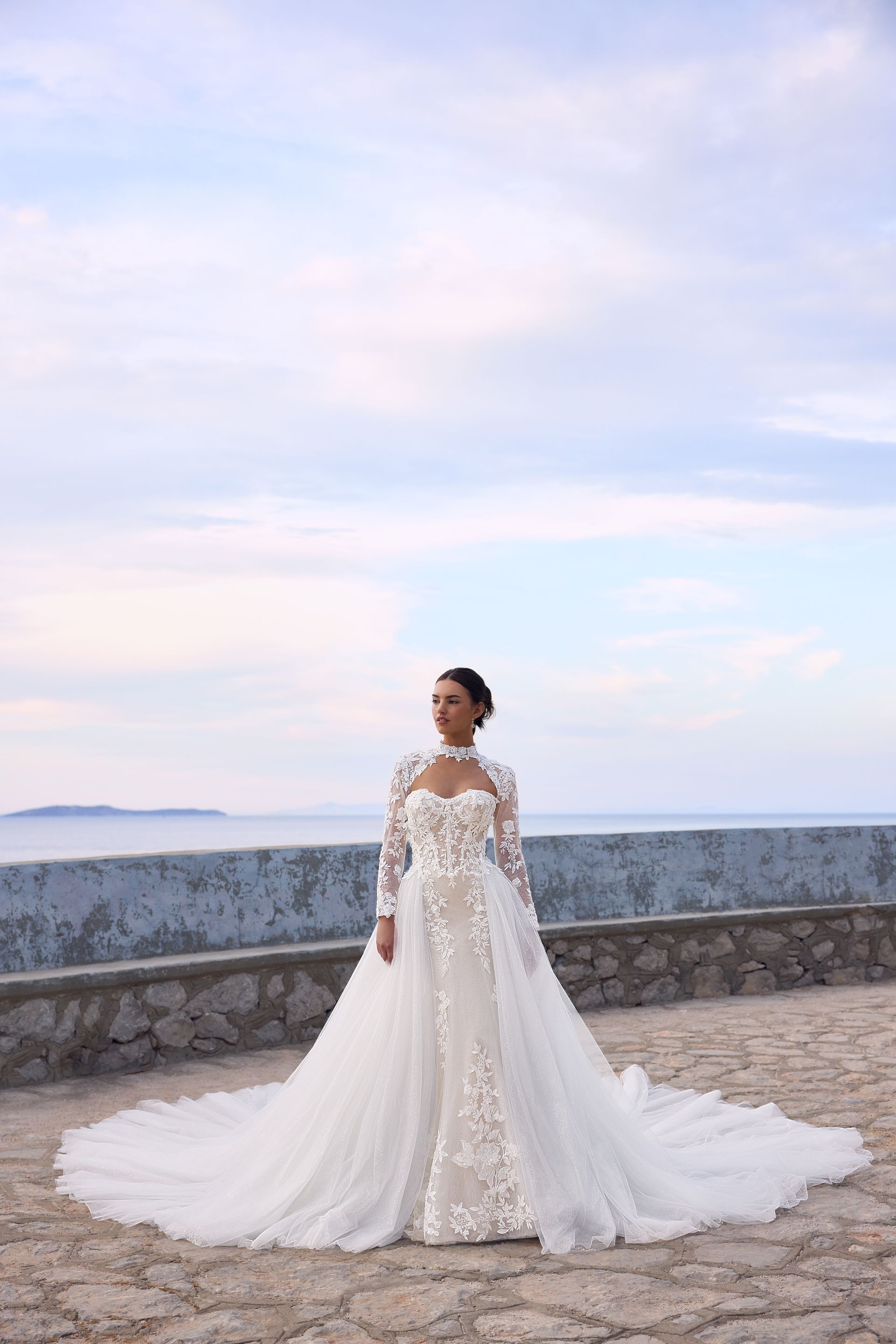 Woman in a white wedding dress with floral embellishments stands by a stone wall, ocean and sky in background.