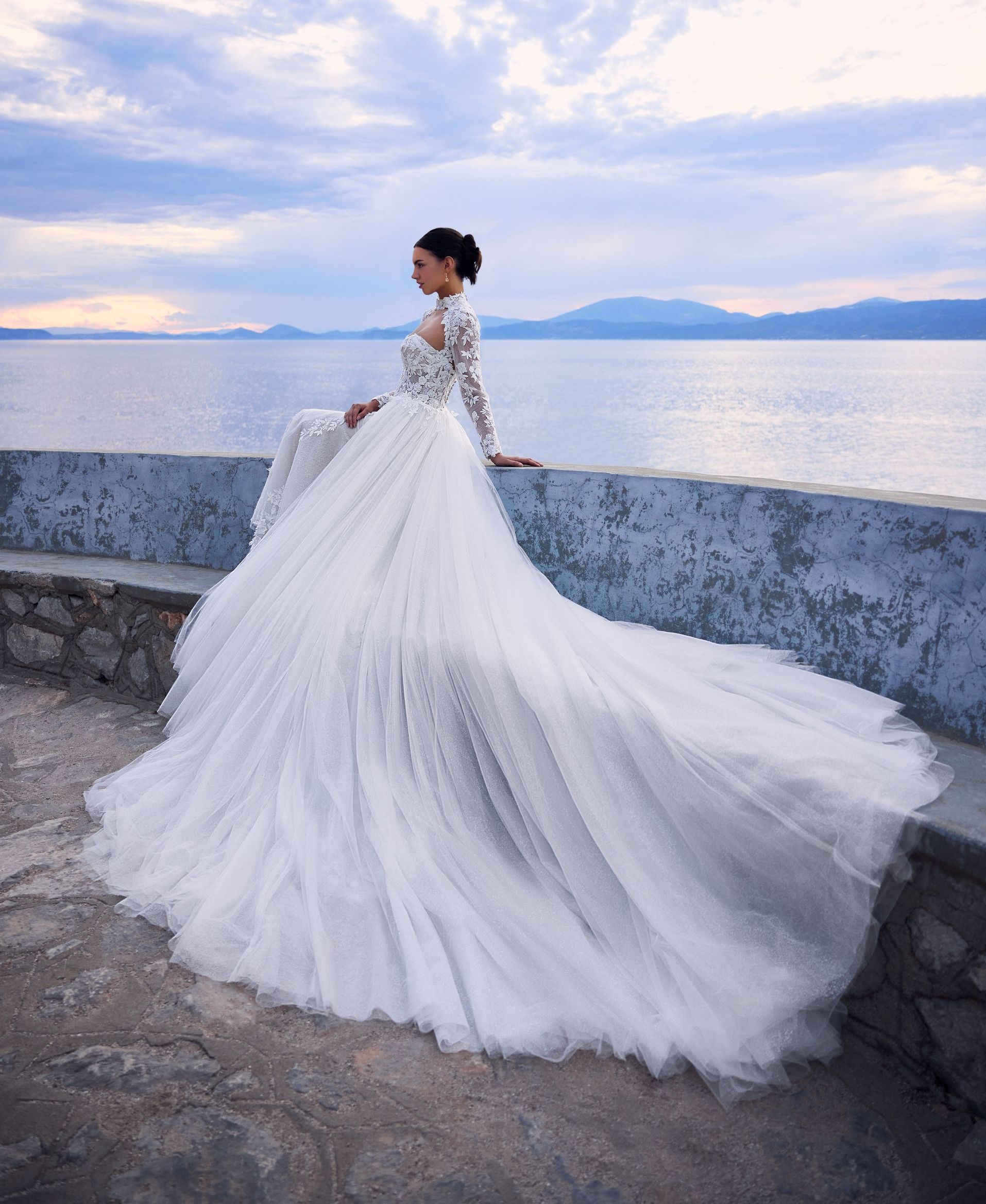Woman in white wedding dress sits on wall overlooking sea.