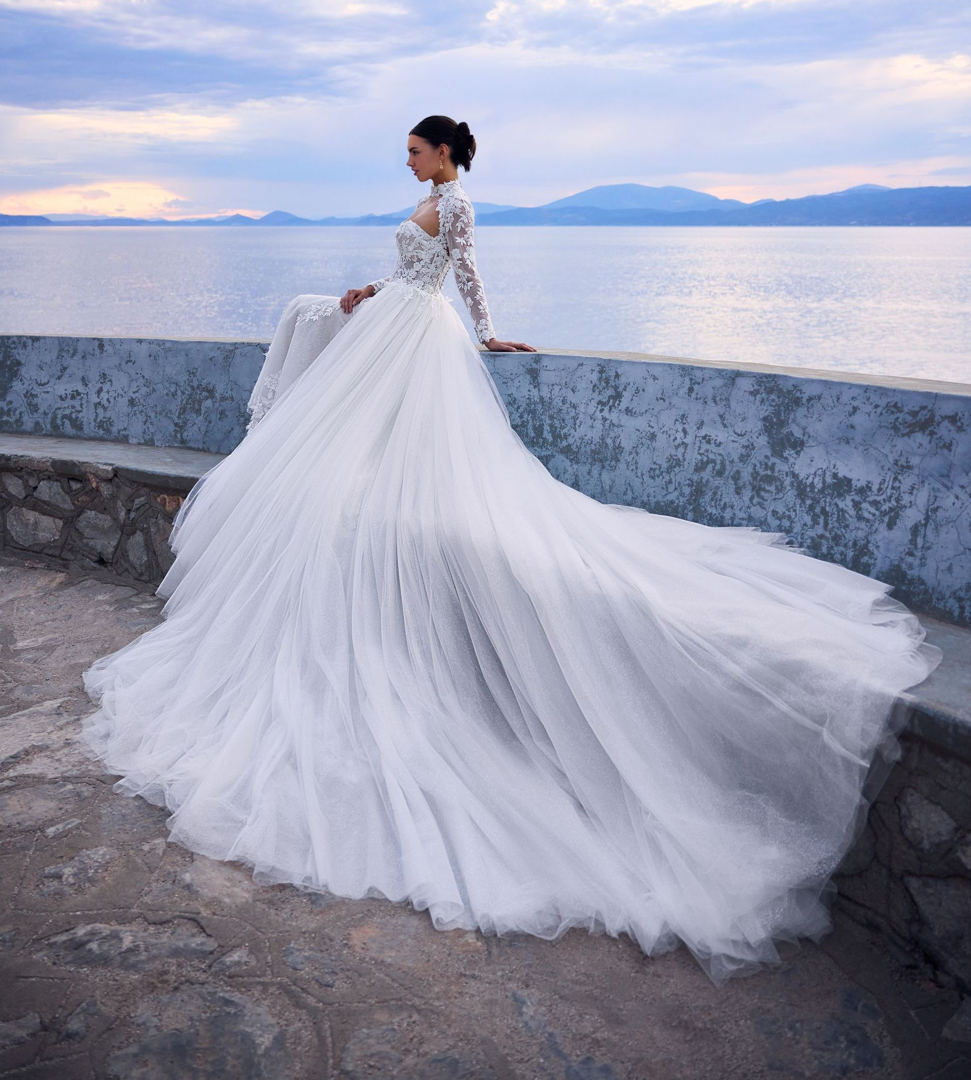 Woman in a long white wedding dress sits on a stone wall, ocean view, cloudy sky.