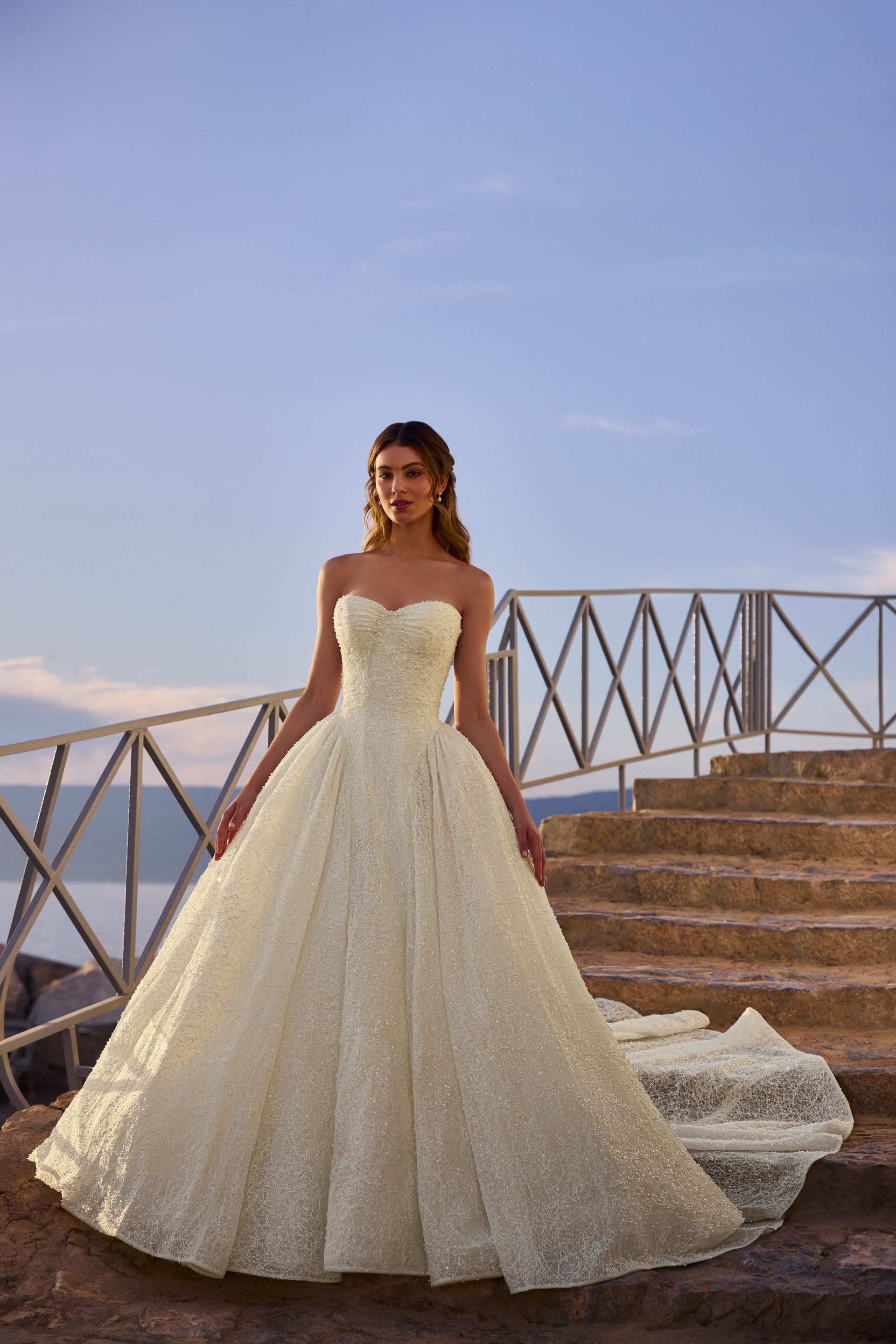 Woman in a beaded strapless gown poses on stone steps with a water view.