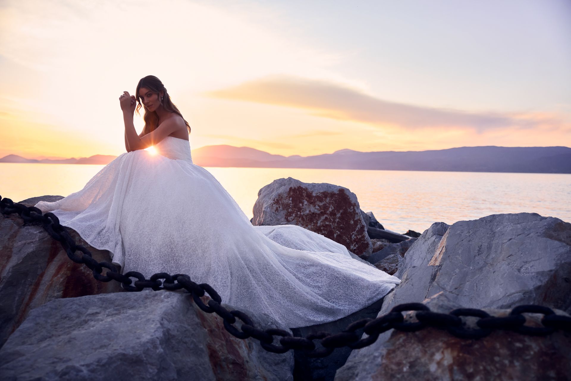 Bride in a white gown sits on rocks, ocean sunset behind her.