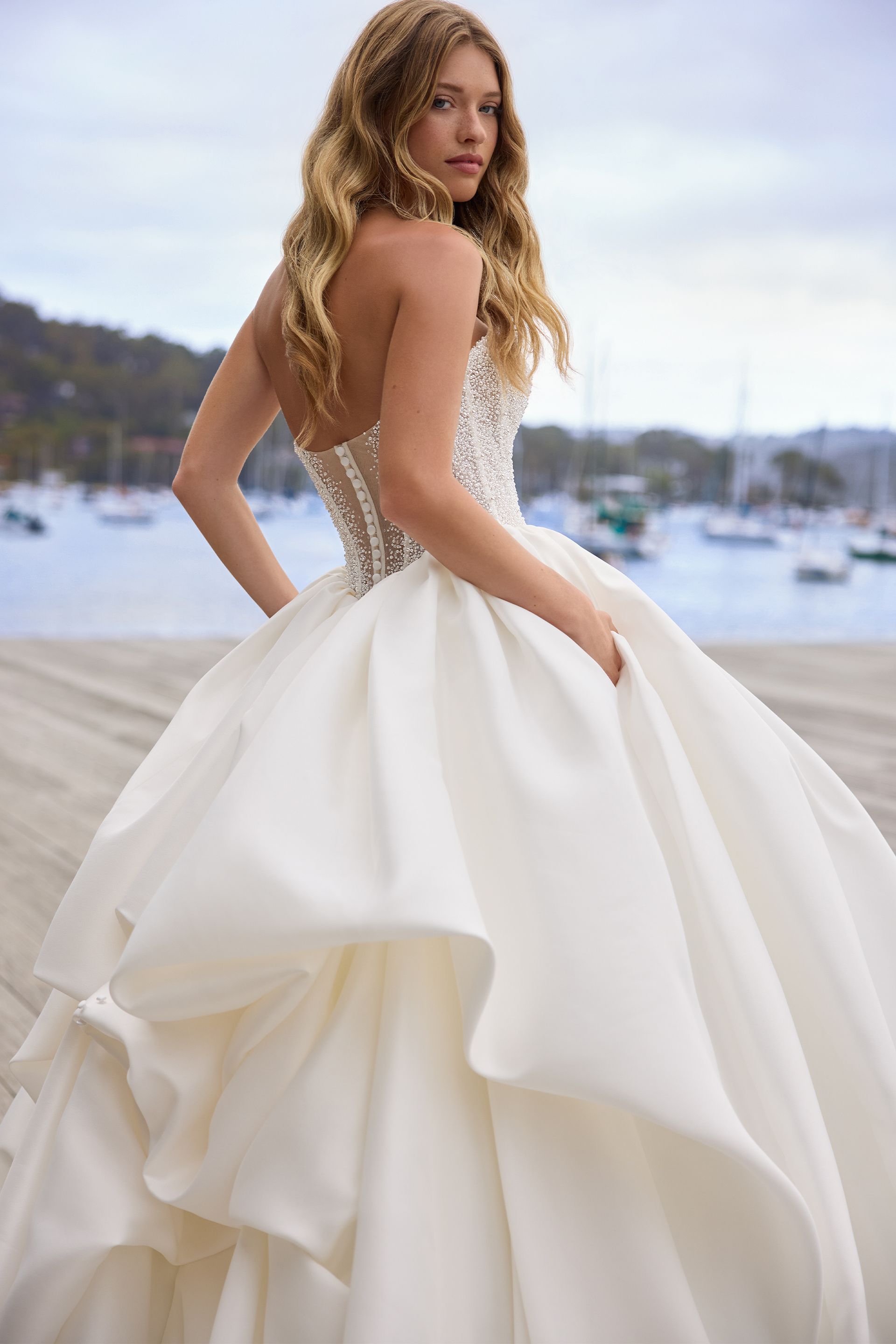 Woman in a white strapless ball gown looking over her shoulder, on a dock, with a harbor in the background.