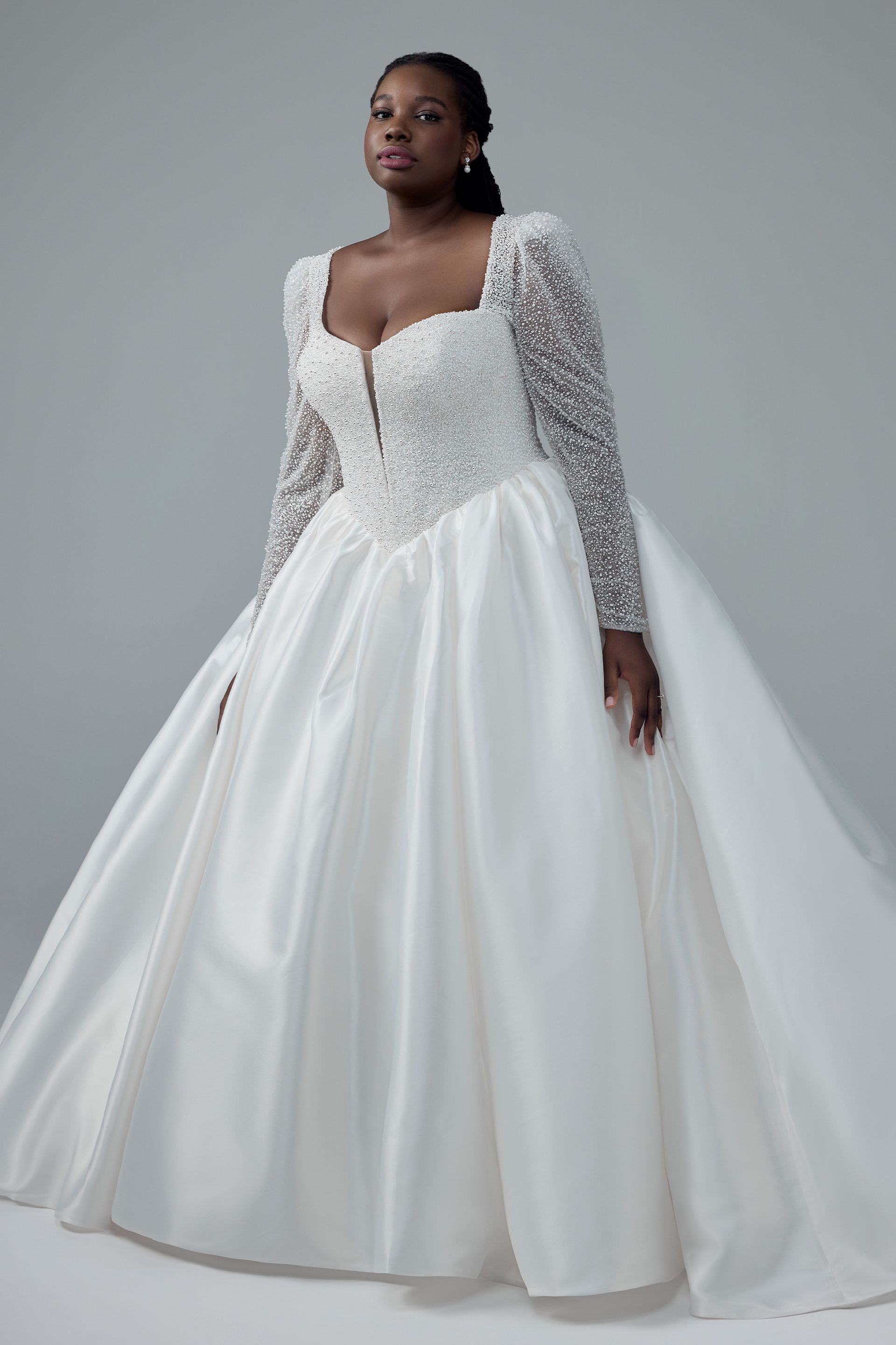 Woman in a white ball gown with long lace sleeves and a square neckline, poses against a light background.