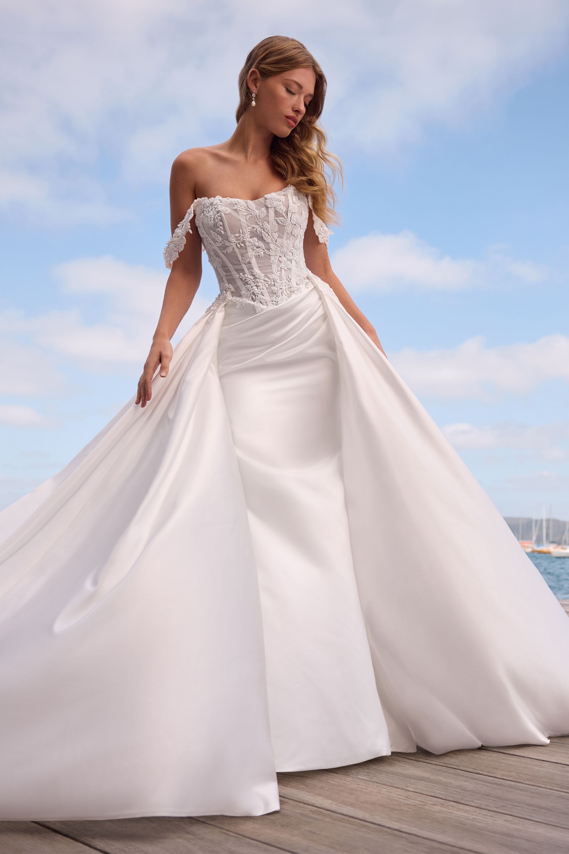 Woman in strapless white wedding dress with detachable skirt, standing on a pier, blue sky background.