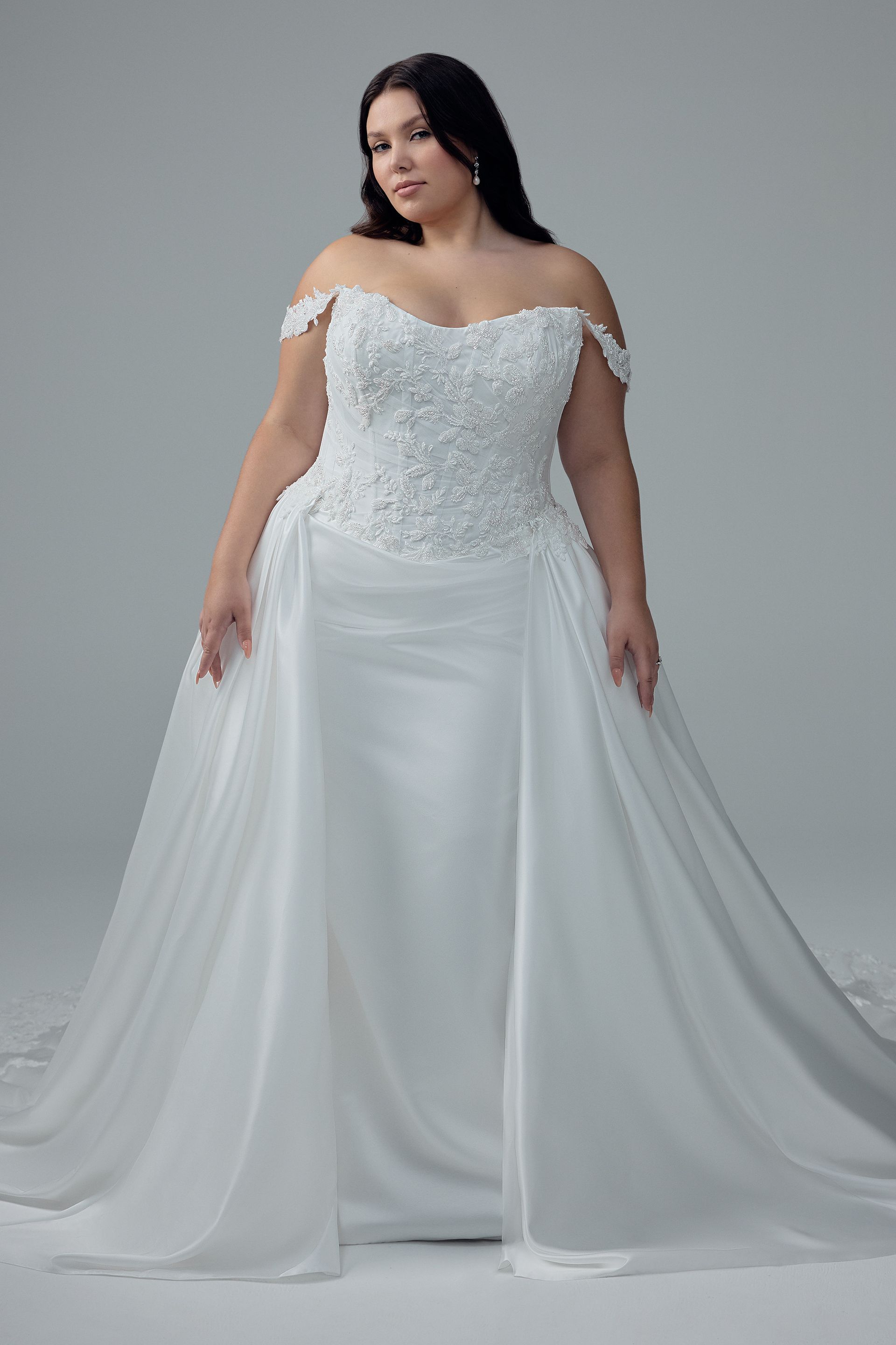 Woman in white off-the-shoulder wedding gown, standing, smiling slightly.