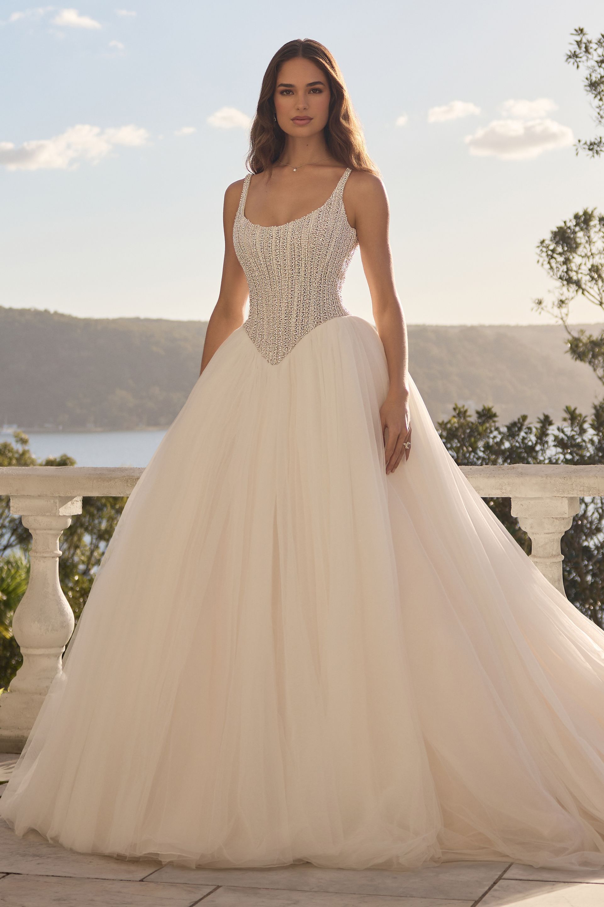 Woman in a white ballgown with jeweled bodice stands outdoors overlooking water.
