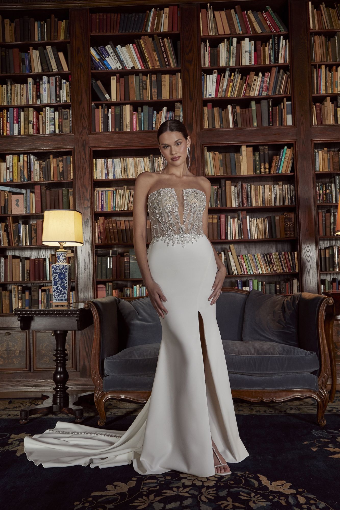 Woman in white gown with bejeweled bodice and thigh slit, standing in a library.