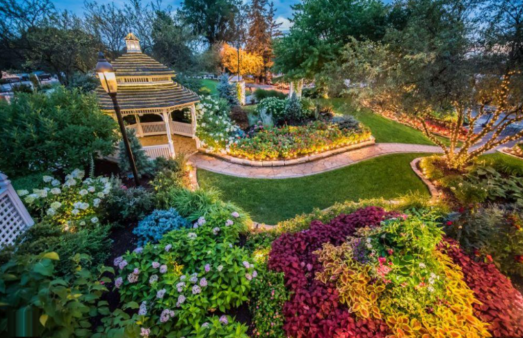 A lush garden scene with a gazebo, paths, and colorful flowers, illuminated by lights at dusk.