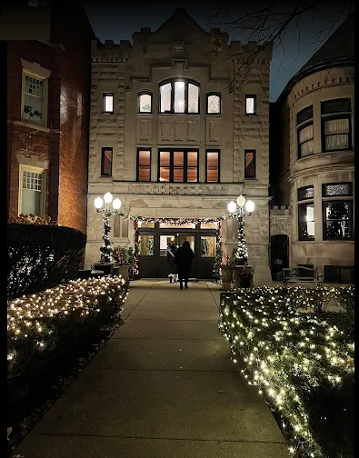 A person walks towards a festively lit, three-story building at night. Lights line the path and bushes.
