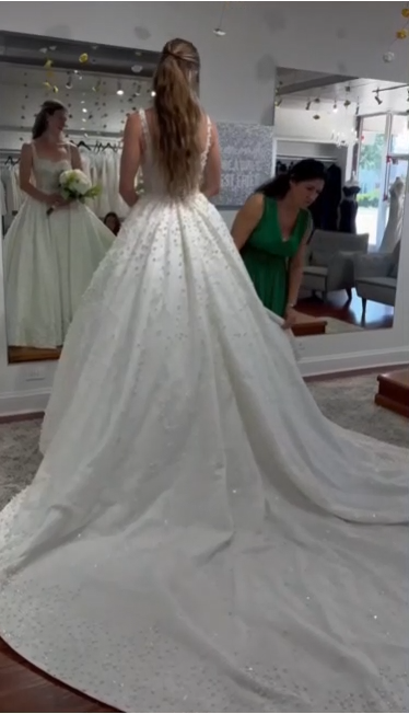 Bride in wedding gown with long train, inside bridal shop. Woman in green dress adjusts the gown. Bride in wedding gown with long train, inside bridal shop. Woman in green dress adjusts the gown.