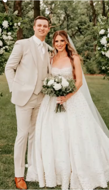 Bride and groom pose outdoors. The bride wears a white gown; the groom wears a tan suit. Bride and groom pose outdoors. The bride wears a white gown; the groom wears a tan suit.