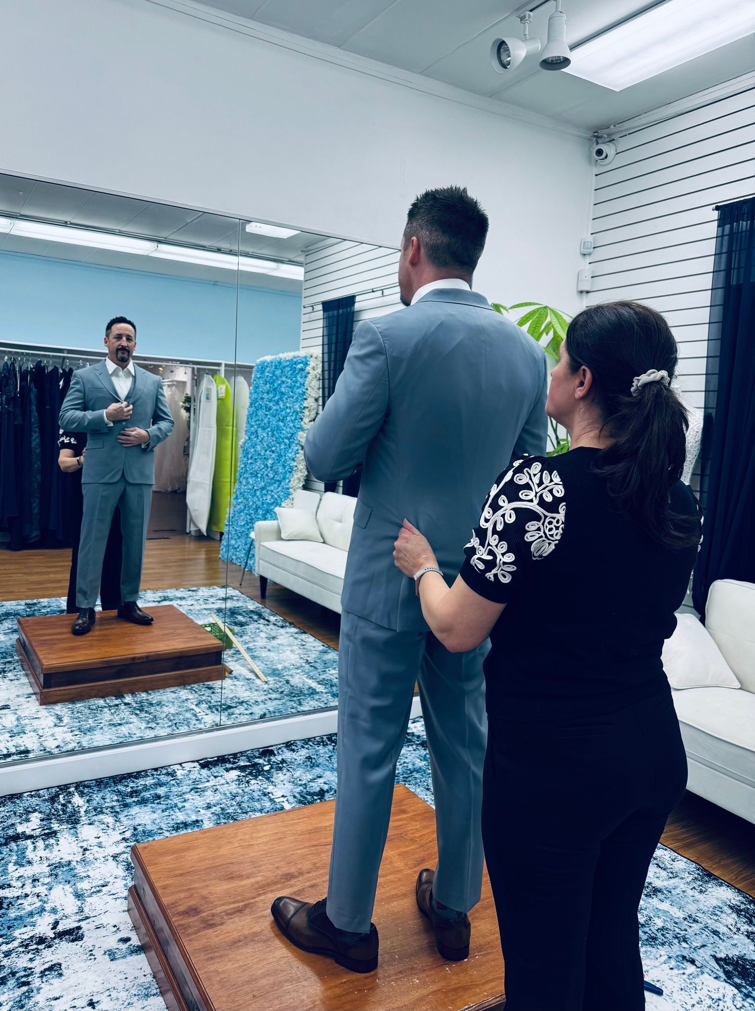 Man in a suit being fitted by a woman in a tailor shop. He stands on a platform in front of a mirror.