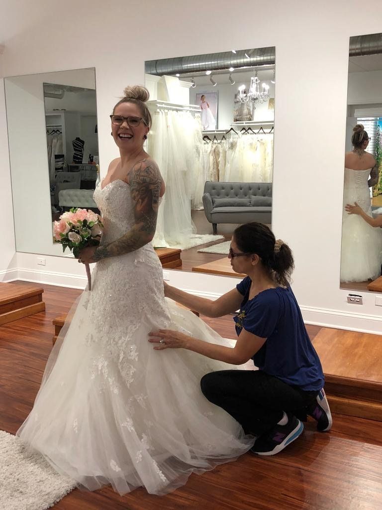 Woman in wedding dress being fitted in a bridal shop. Assistant adjusts the gown's skirt.
