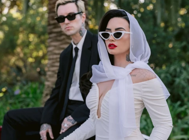 Woman in white dress and veil with man in black suit, both wearing sunglasses, seated outdoors.