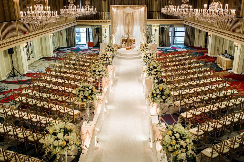 Wedding ceremony setup in grand ballroom, rows of chairs face an altar, white aisle runner, floral arrangements.