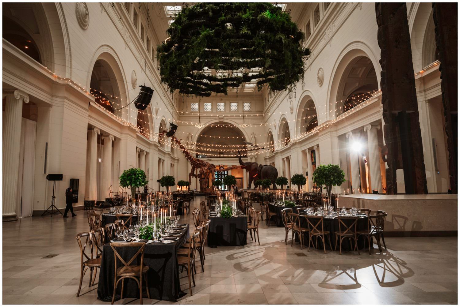 A banquet hall set for an event with round and rectangular tables, a large floral installation, and dinosaur skeletons.