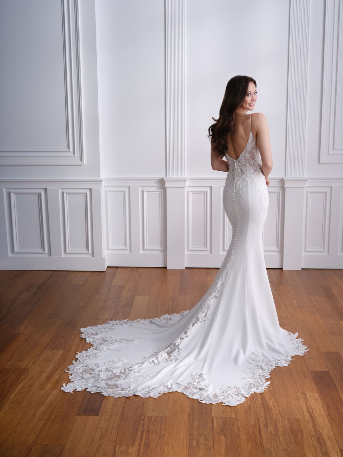 Woman in a white lace gown standing against a paneled wall, hands clasped and facing forward.