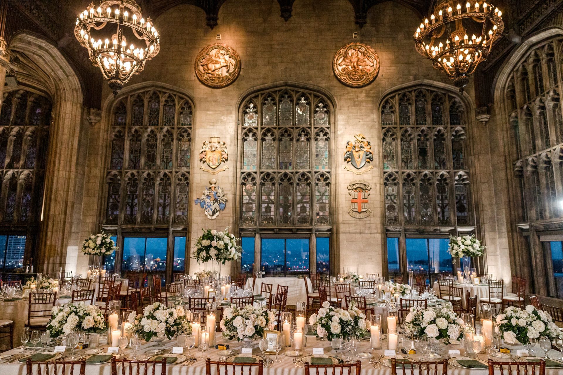 Elegant banquet hall with tall arched windows, tables set for a formal event, and city skyline view.