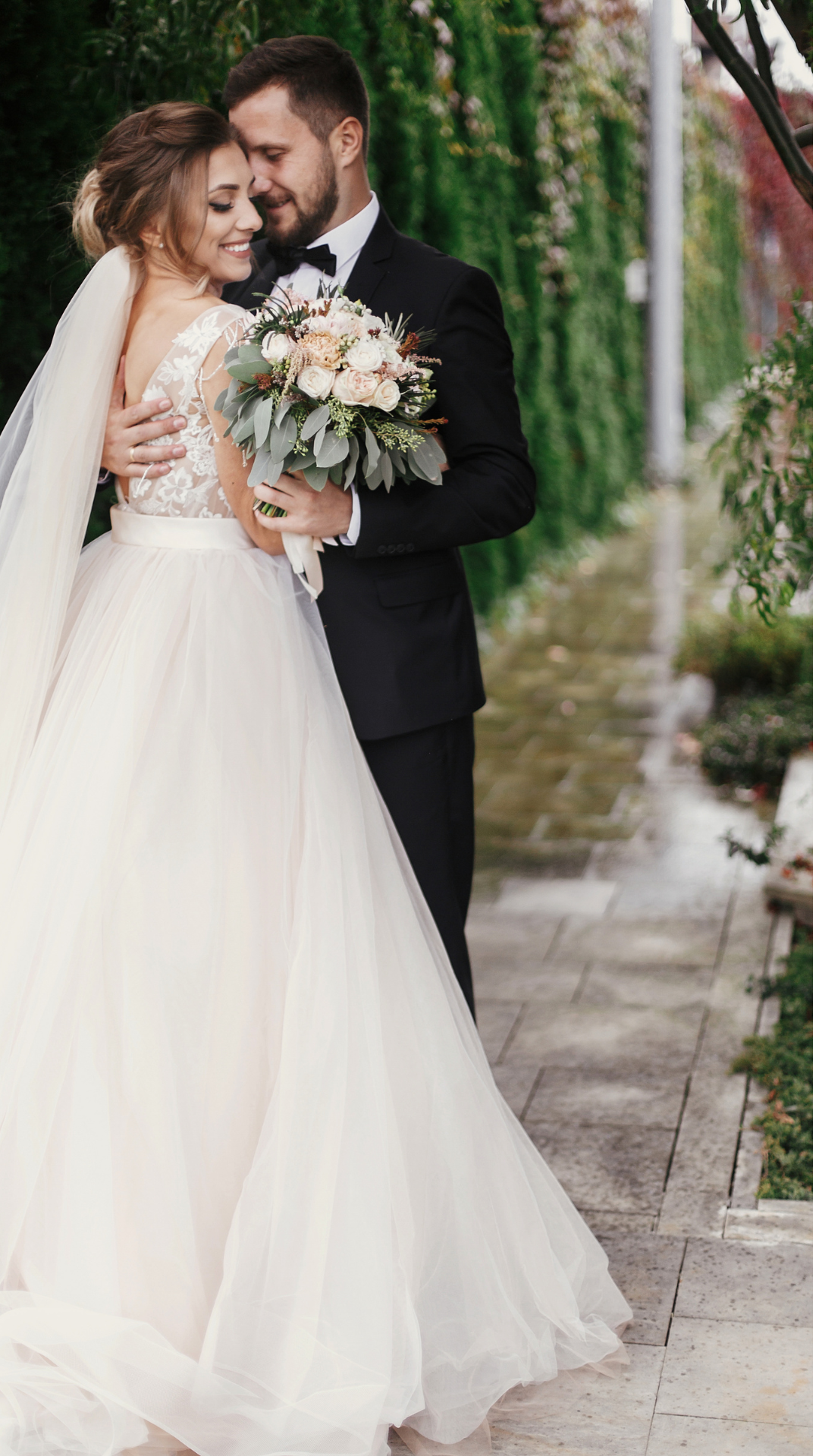 Bride and groom embrace outdoors; bride in white gown with bouquet, groom in black tuxedo.