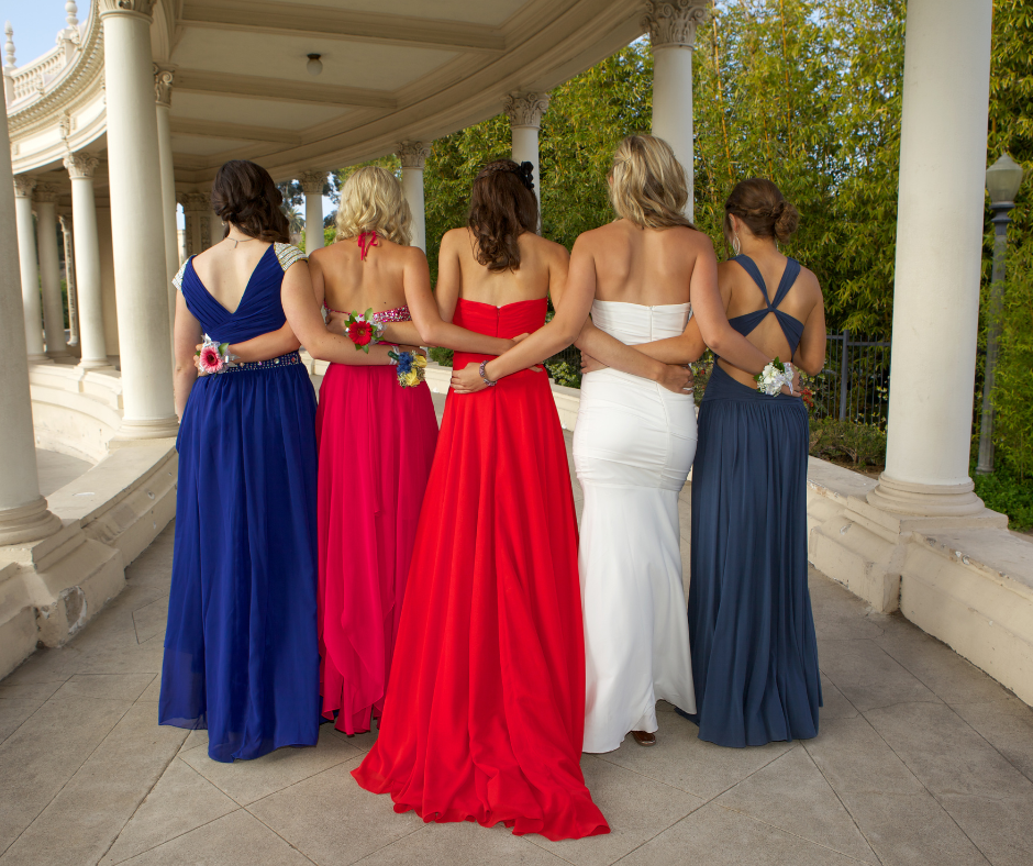 Five women in prom dresses stand with arms around each other under a columned walkway.