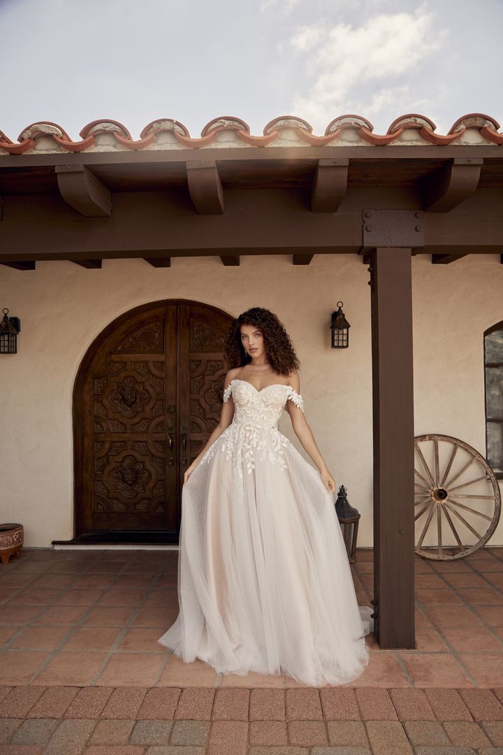 Woman in a white off-the-shoulder wedding dress stands in front of a carved wooden door.