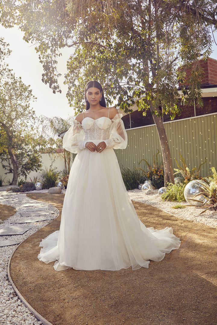 Woman in white wedding dress with off-the-shoulder sleeves stands outdoors, holding hands.