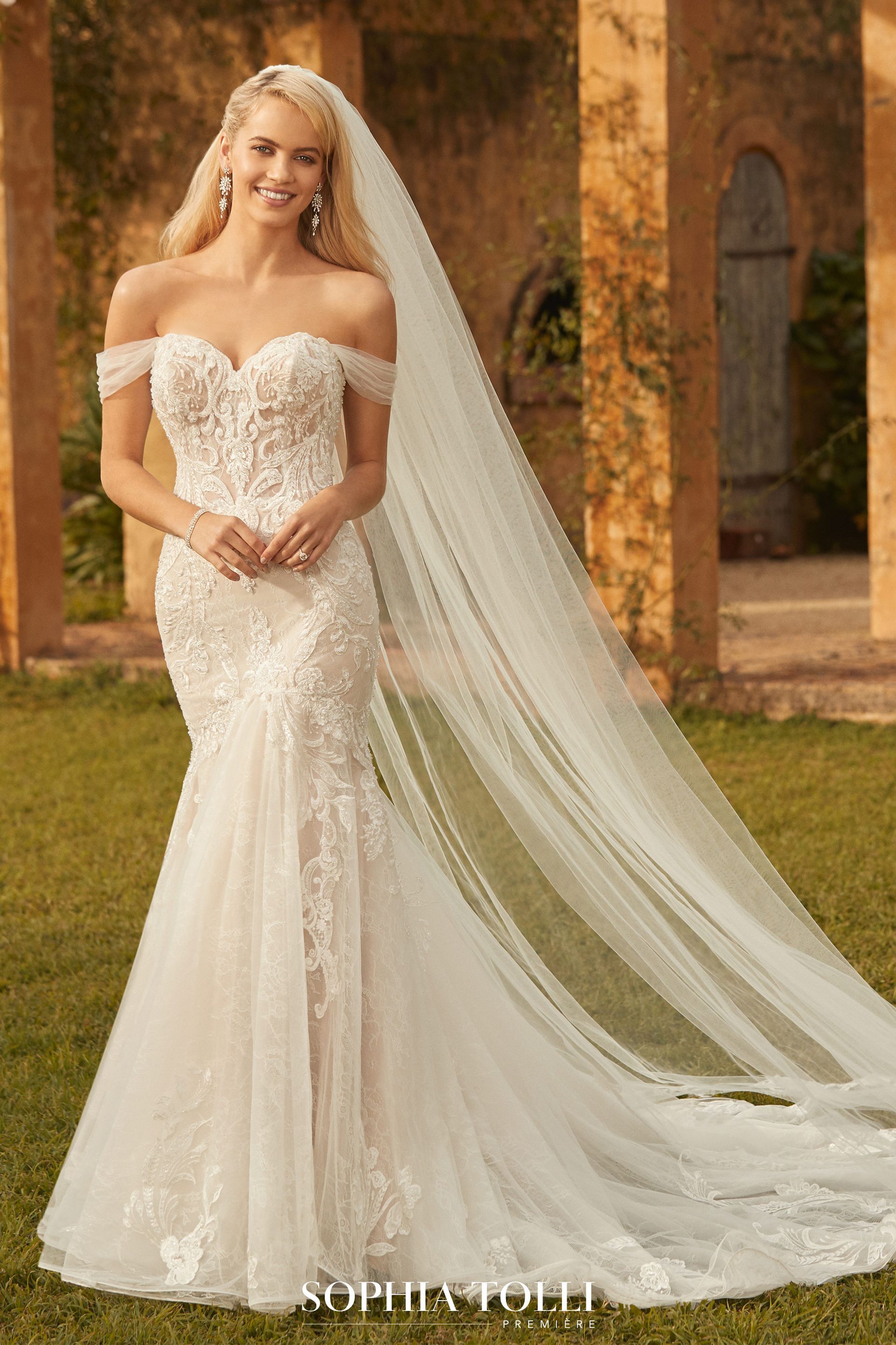 Woman in a white, off-the-shoulder wedding dress with a long veil in an outdoor setting.