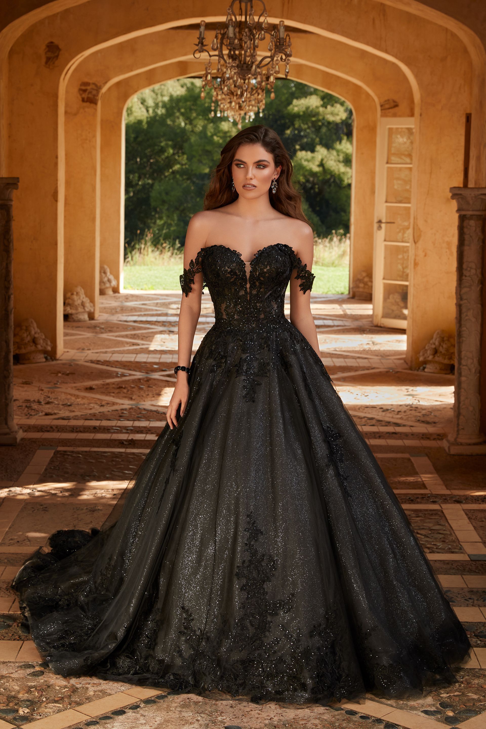 Woman in black off-the-shoulder gown stands in an arched stone corridor; embellished bodice, full skirt.