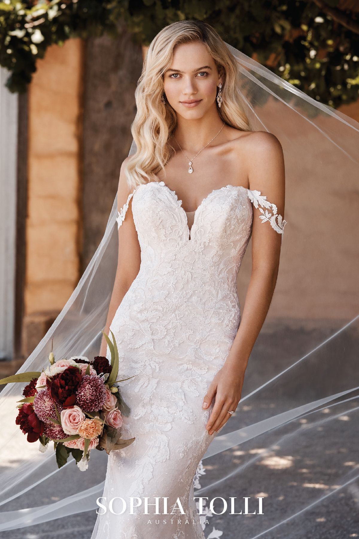 Bride in a lace, off-the-shoulder wedding dress, holding a bouquet, with a veil outdoors.