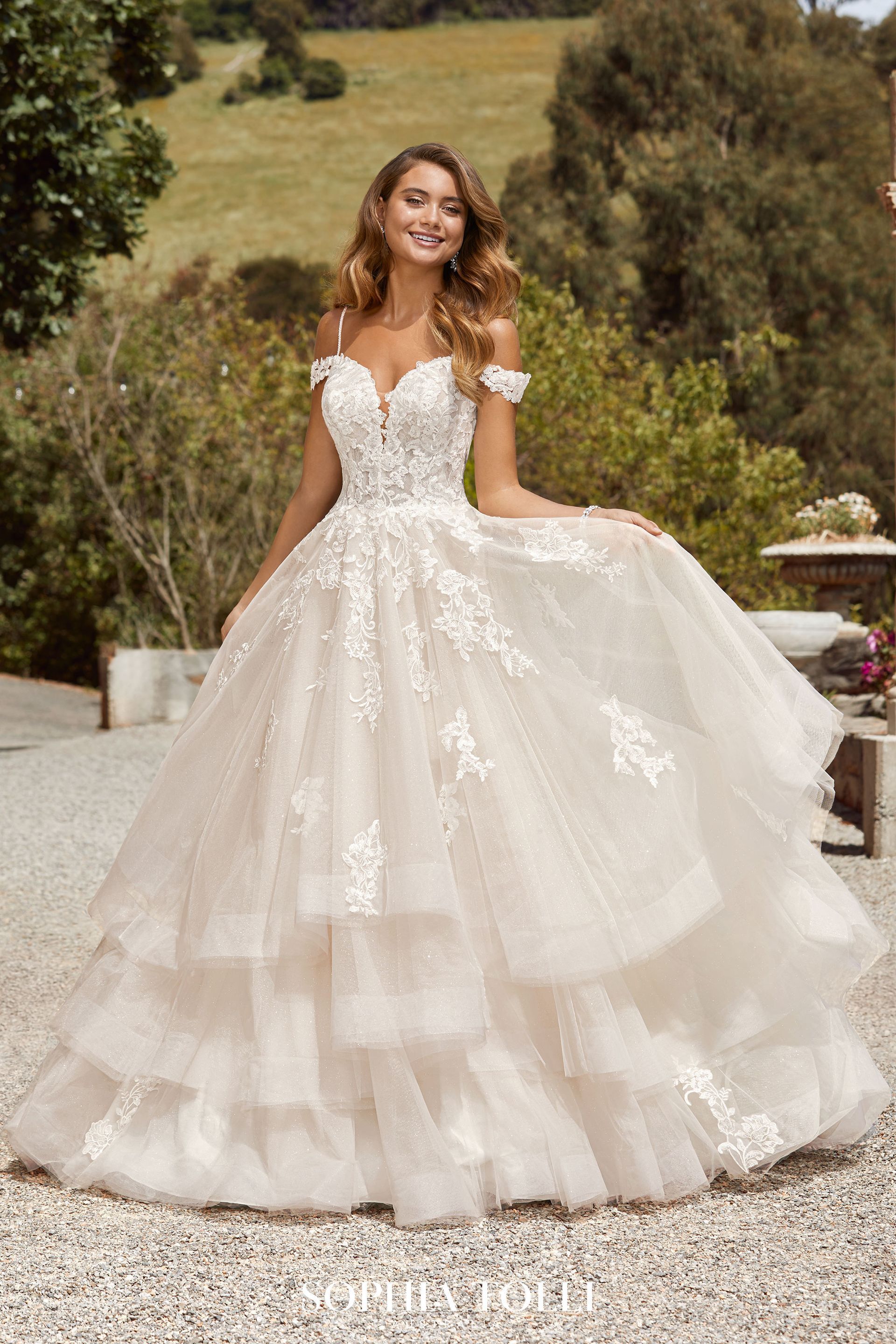 Woman in off-the-shoulder, tiered wedding dress outdoors, smiling, holding skirt.