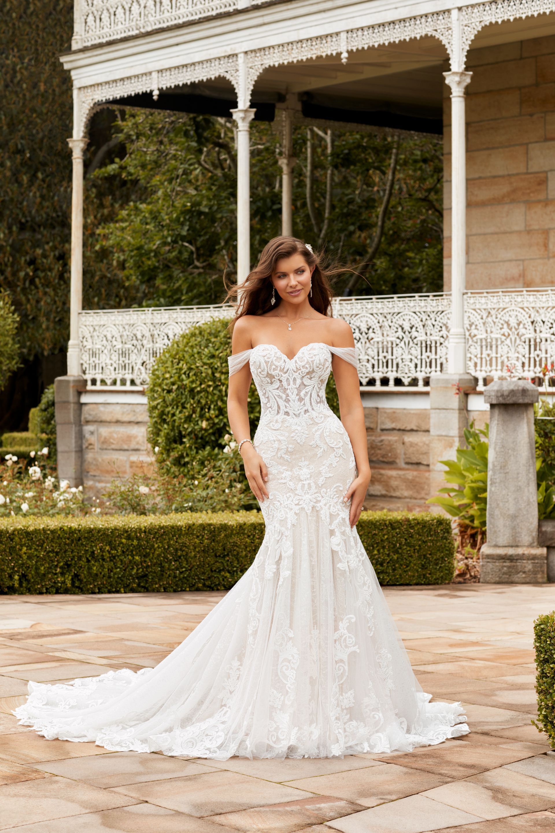 Woman in white off-shoulder lace wedding dress, standing outdoors in front of a house.