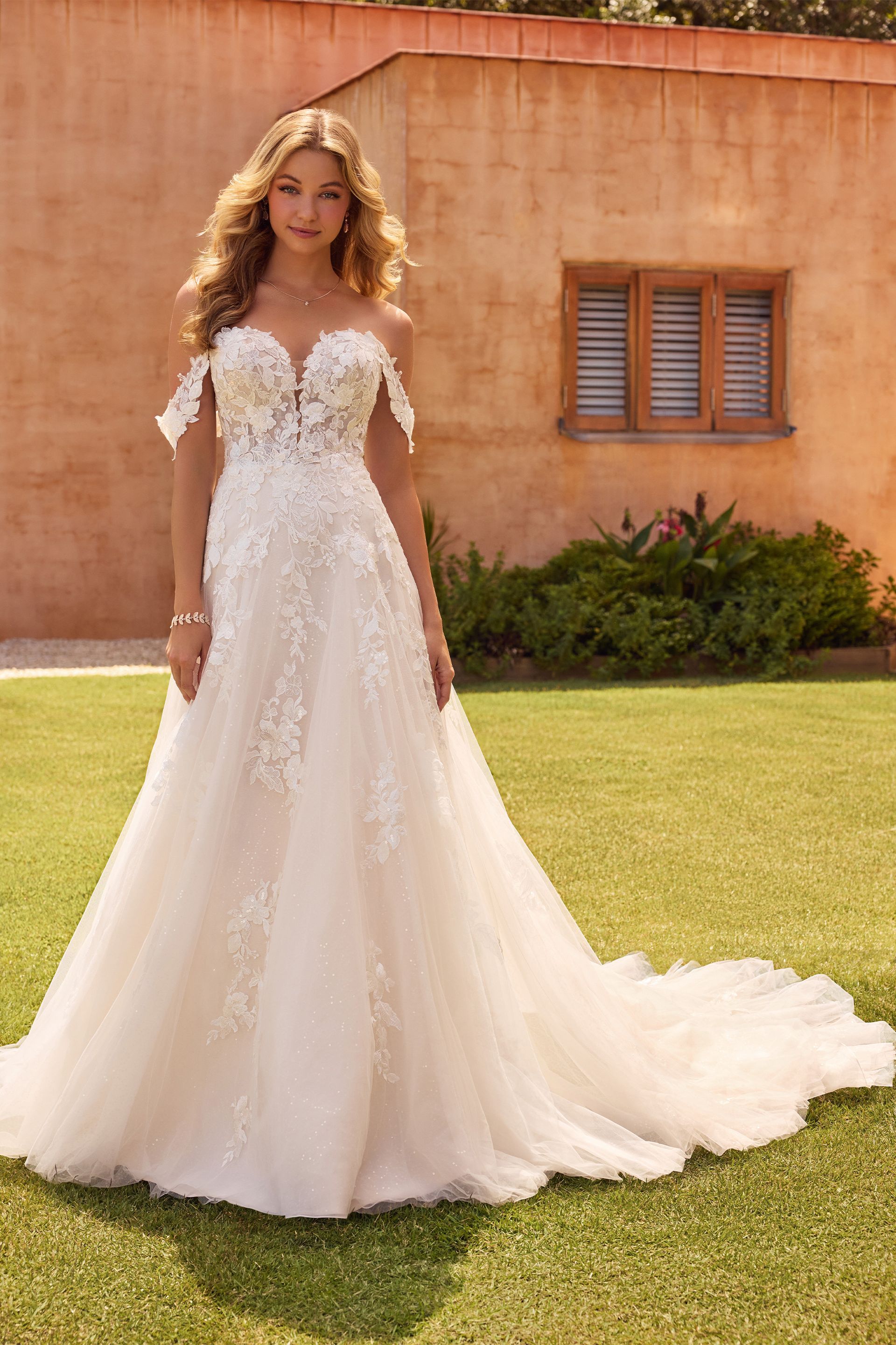 Woman in off-the-shoulder wedding dress with floral embellishments, standing on grass.