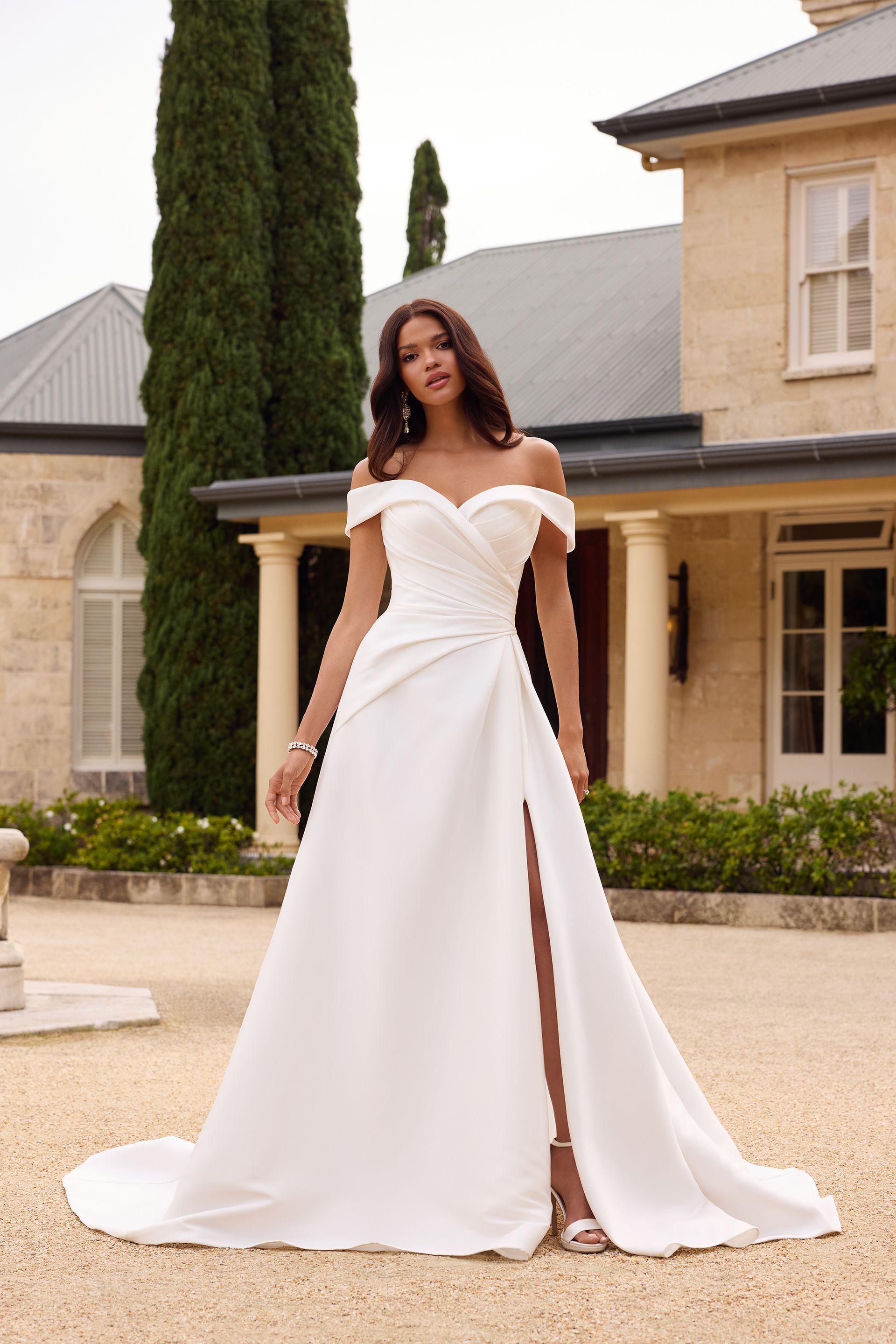 Woman in white off-the-shoulder wedding dress with high slit, standing outdoors near a stone building.