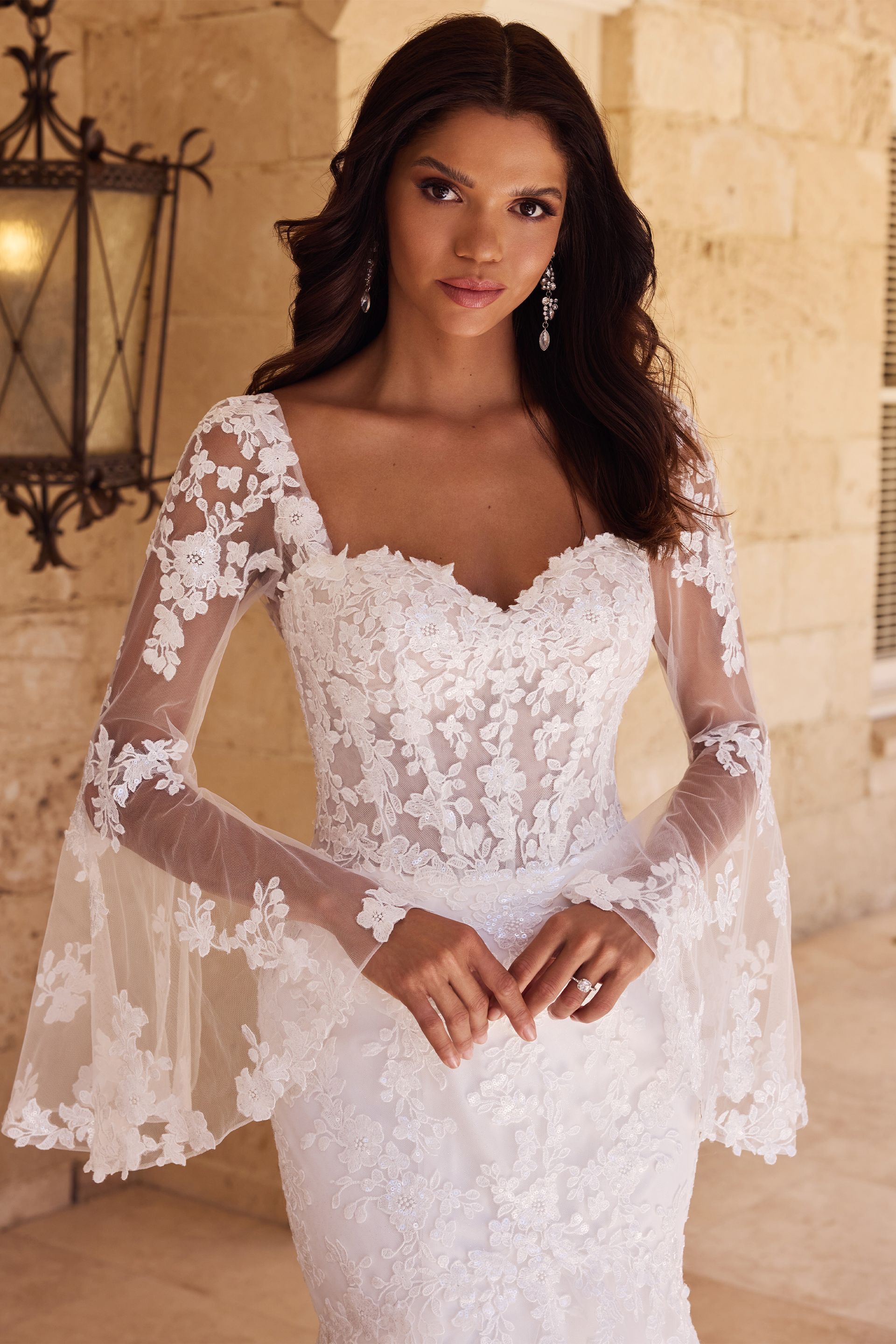 Woman in a white lace wedding dress with flared sleeves, standing near an outdoor wall.
