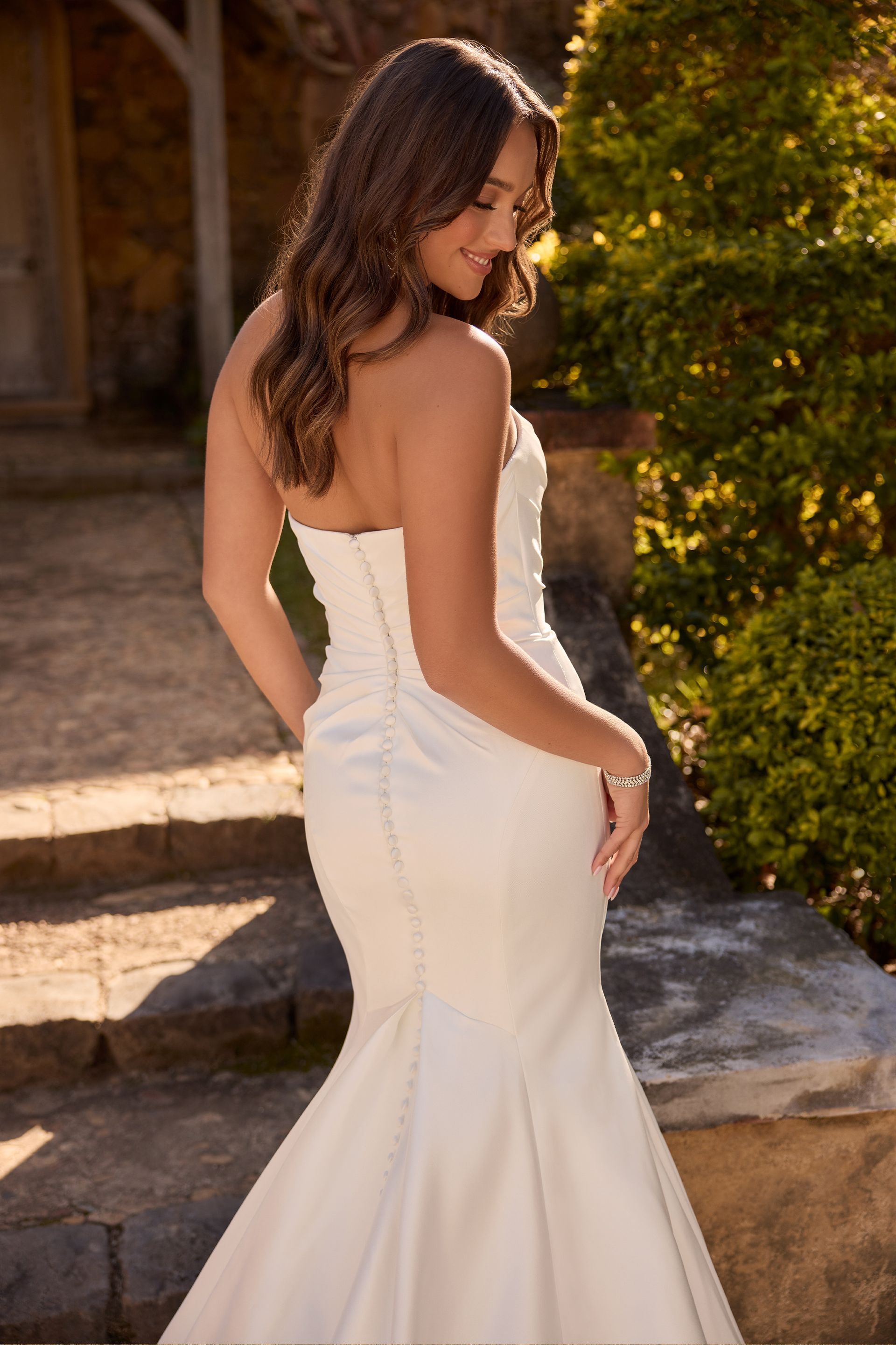 Woman in white strapless dress, looking down, standing on stone steps outdoors, near greenery.