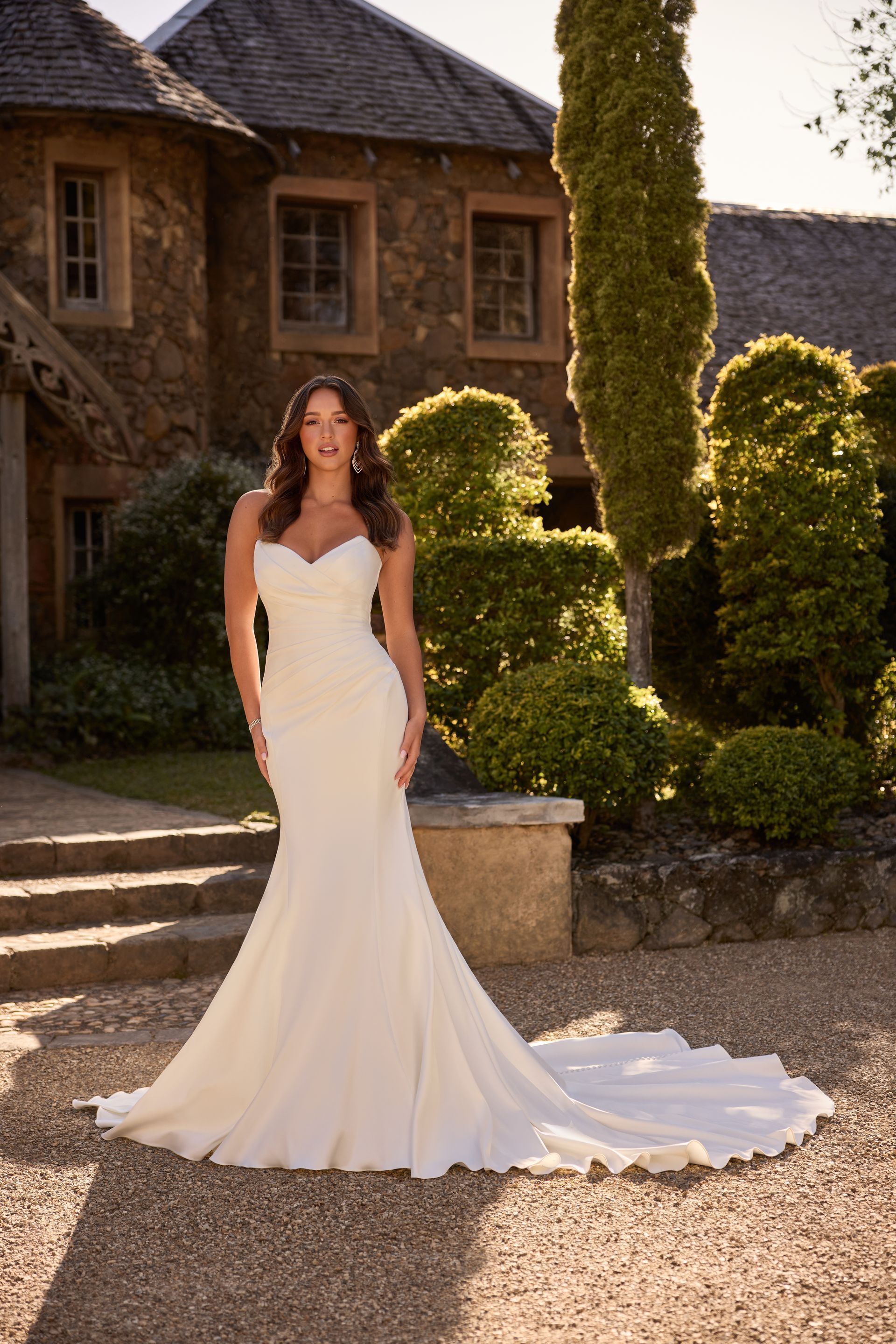 Woman in white wedding dress with train poses outside a stone building.
