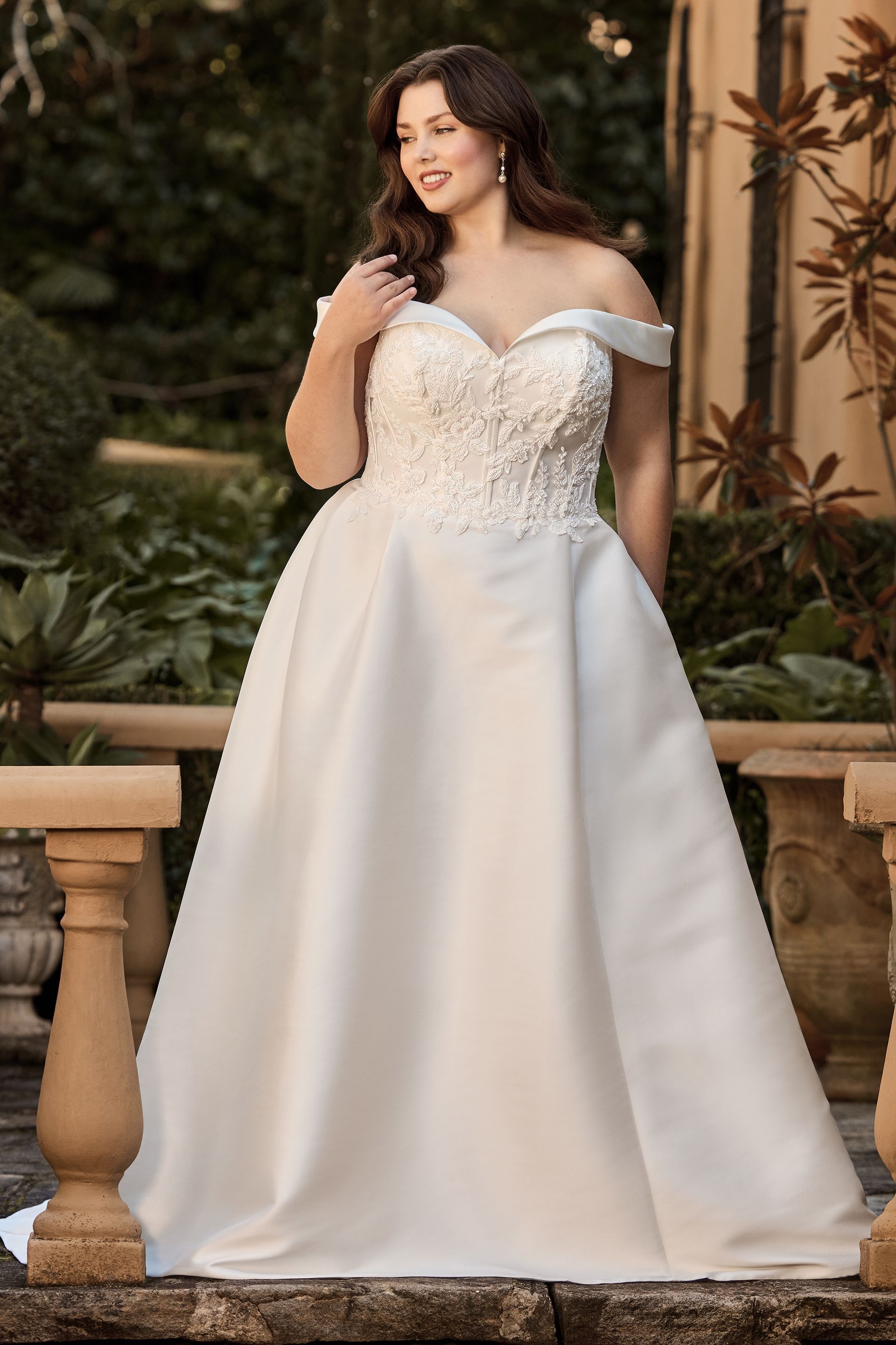 Woman in white off-the-shoulder wedding gown, standing outdoors, posing.