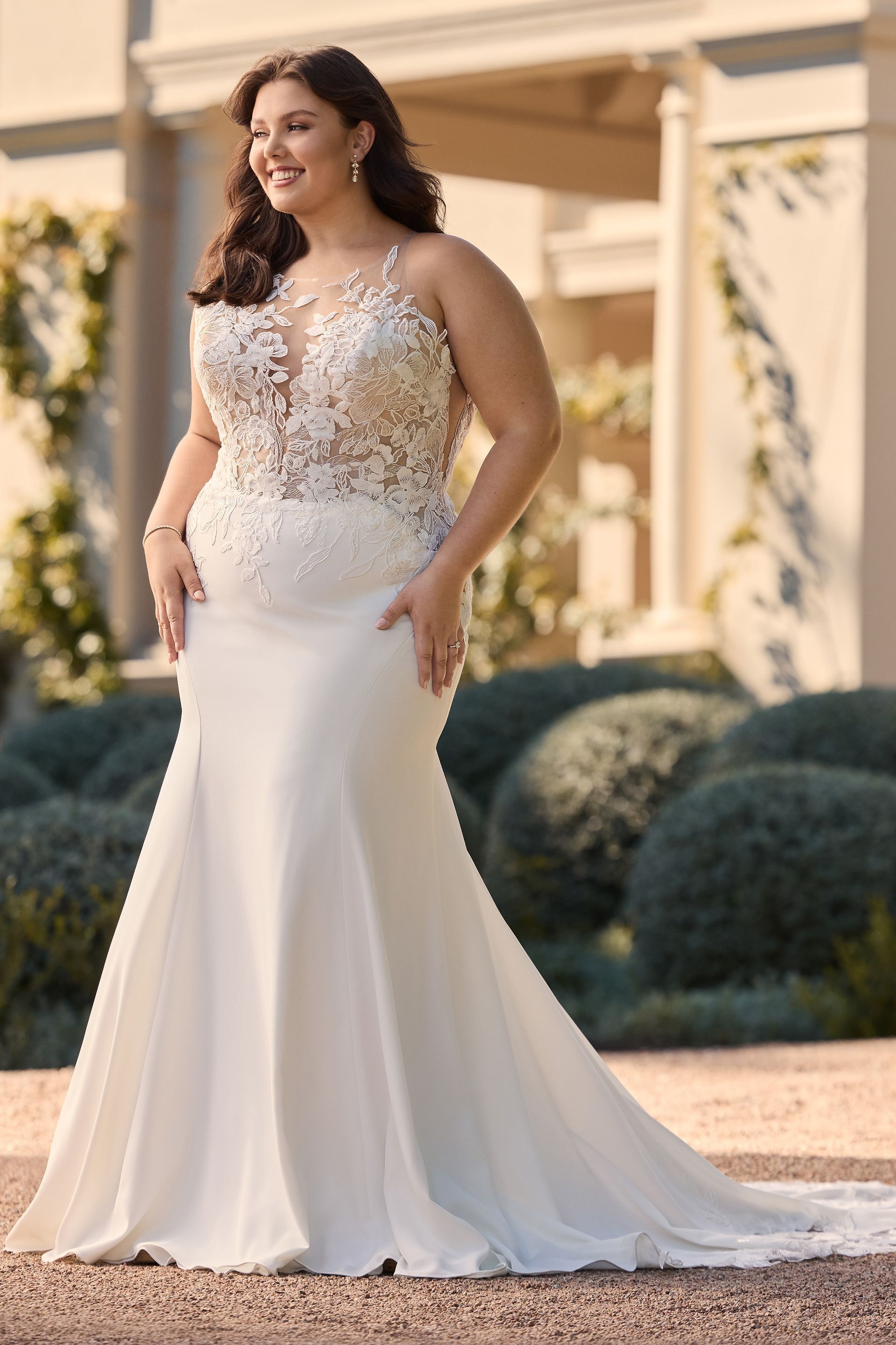 Woman in a white wedding dress smiles outdoors. The dress has lace bodice and a mermaid silhouette.