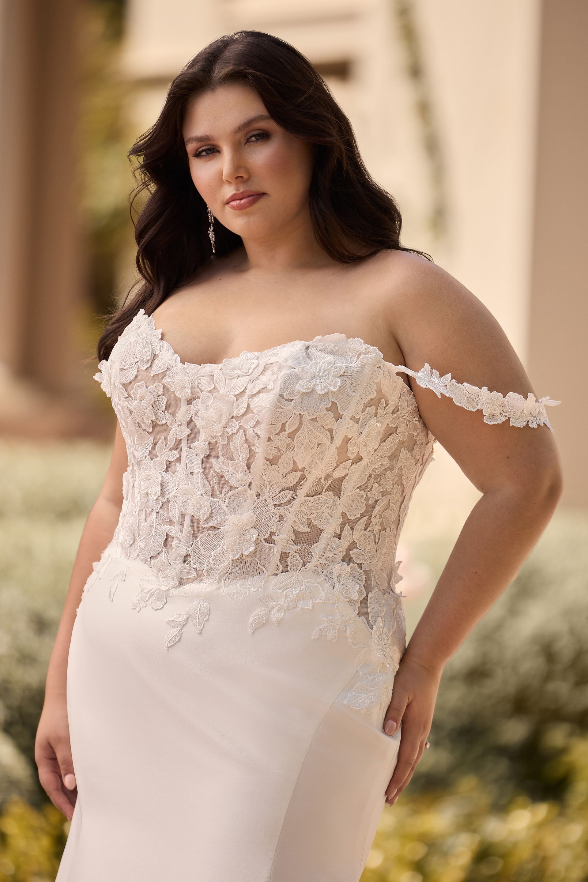 Woman in a white off-the-shoulder wedding dress with lace bodice, looking at the camera. Outdoor setting.