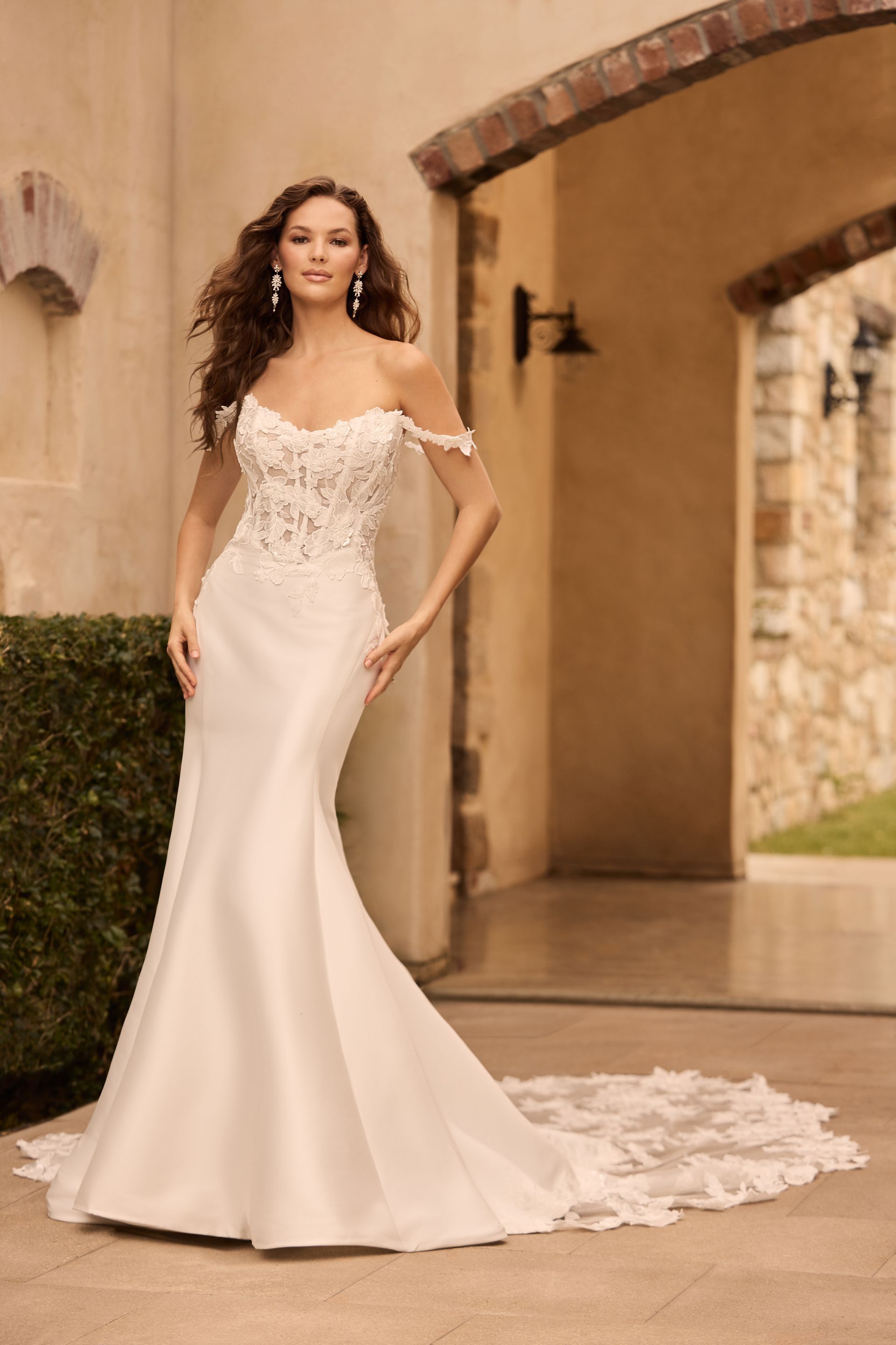 Woman in a white off-the-shoulder wedding dress with a long train, standing in front of an arched doorway.