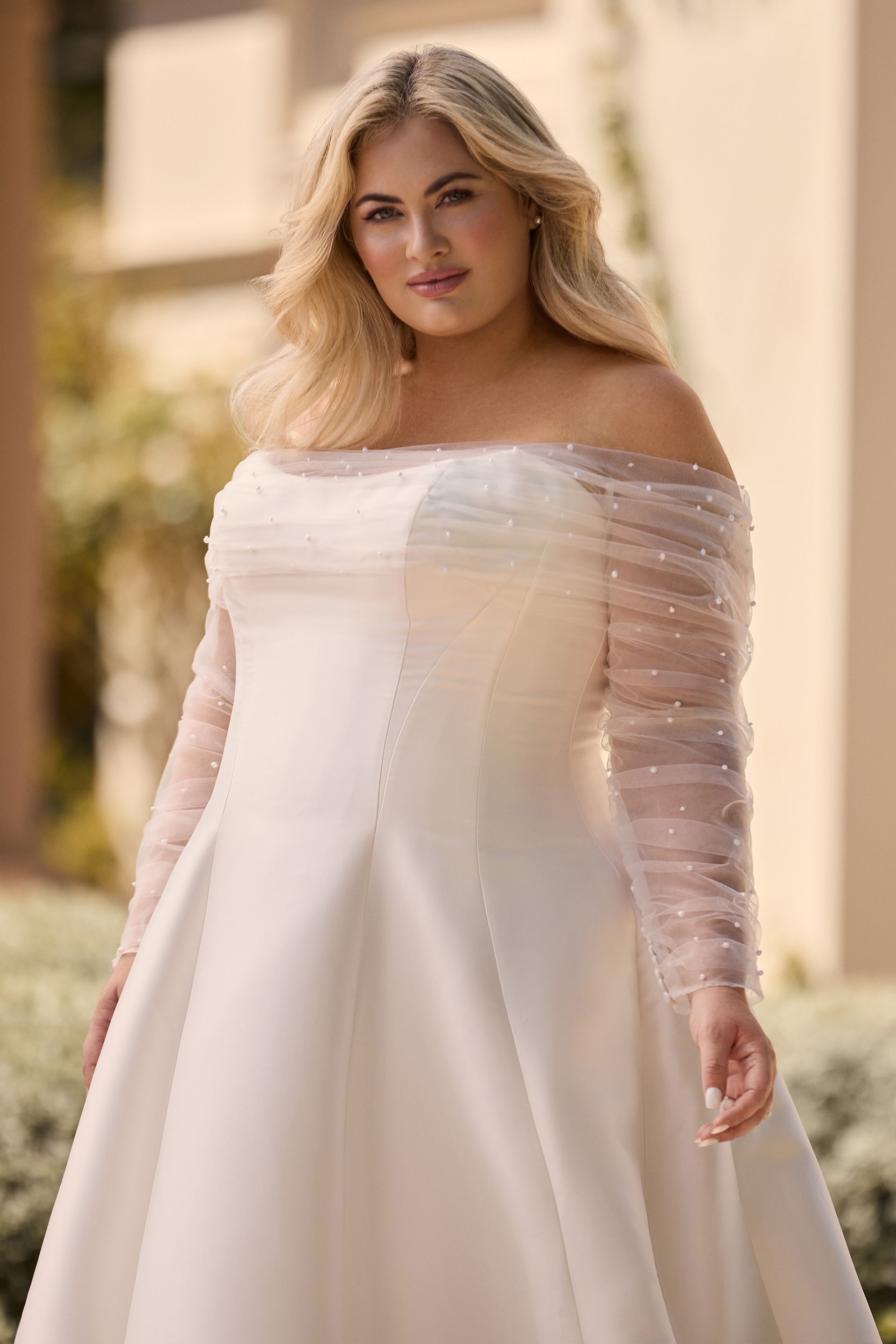 Woman in off-the-shoulder wedding dress with beaded sleeves. Outdoors, natural light, smiling.