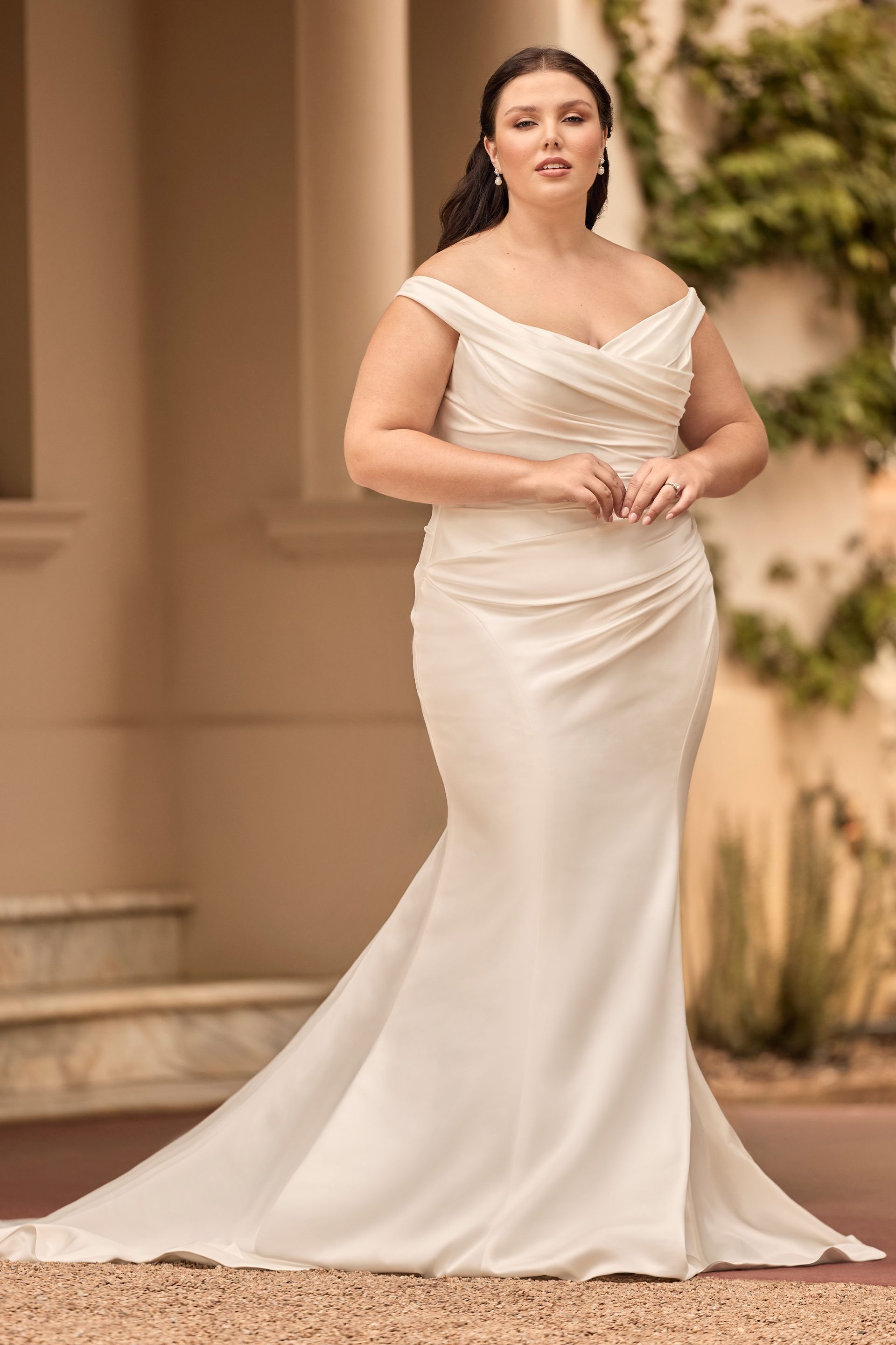 Woman in off-the-shoulder ivory wedding dress with a train, posing outdoors.