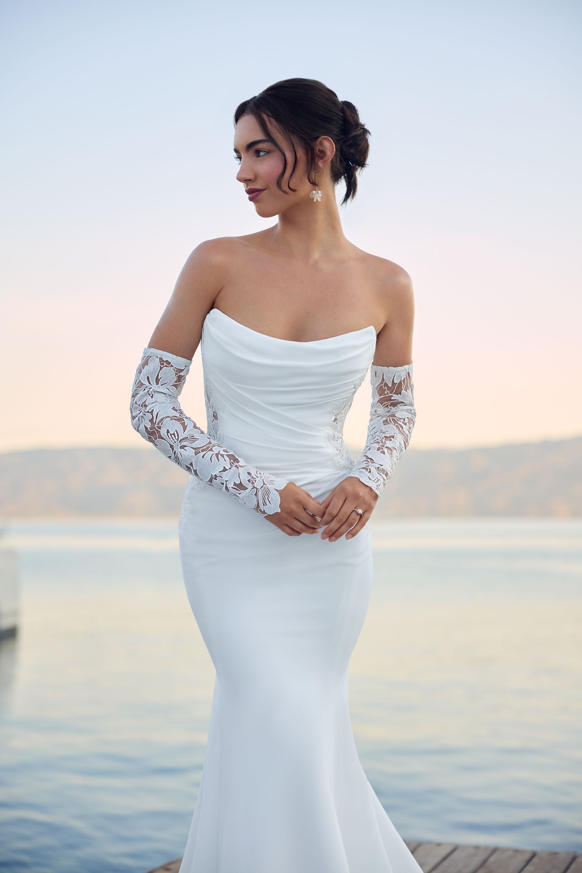 Woman in a white strapless gown with lace sleeves poses by water.