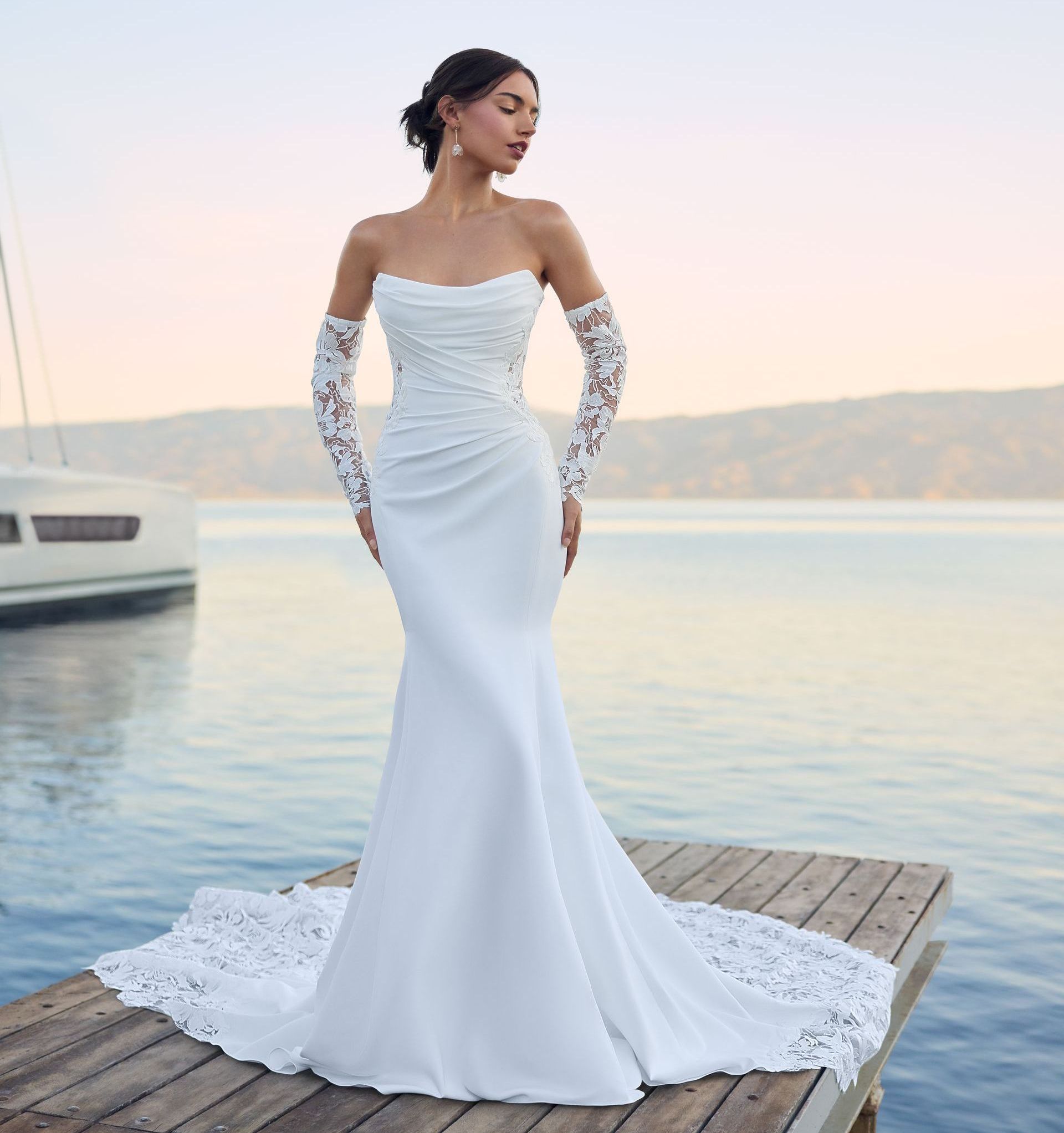 Woman in a white wedding dress with lace sleeves and a train, standing on a dock by water.