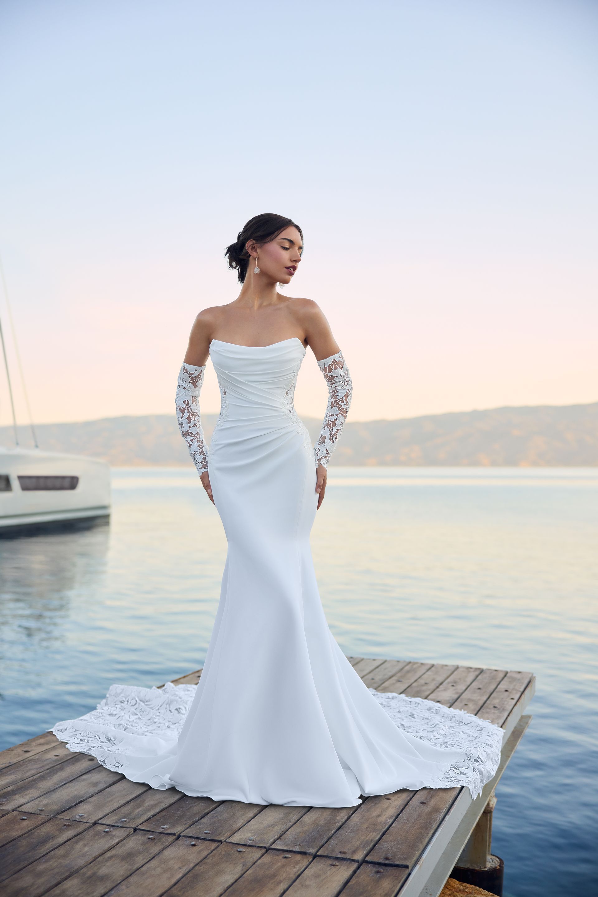 Woman in strapless white wedding dress with lace gloves and train, standing on a dock by water.