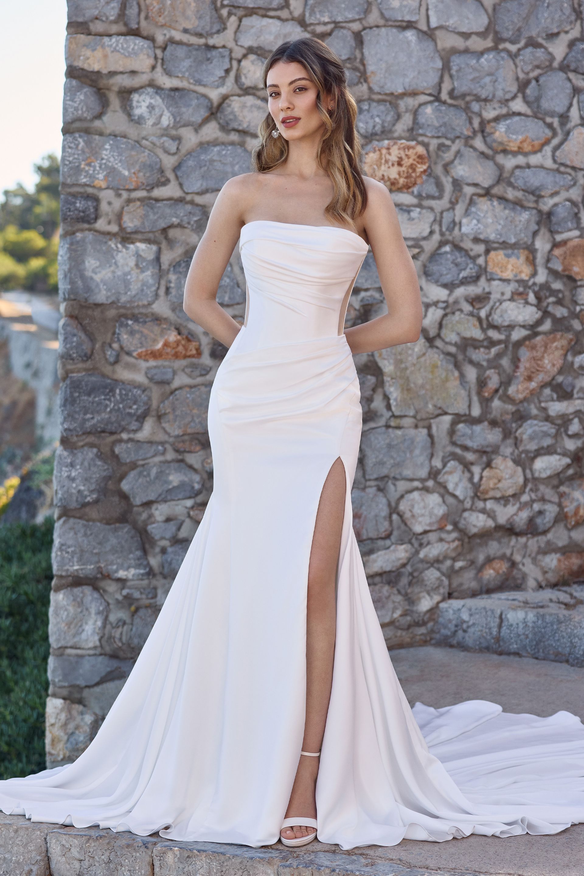 Woman in strapless white wedding dress with high slit, standing against stone wall.