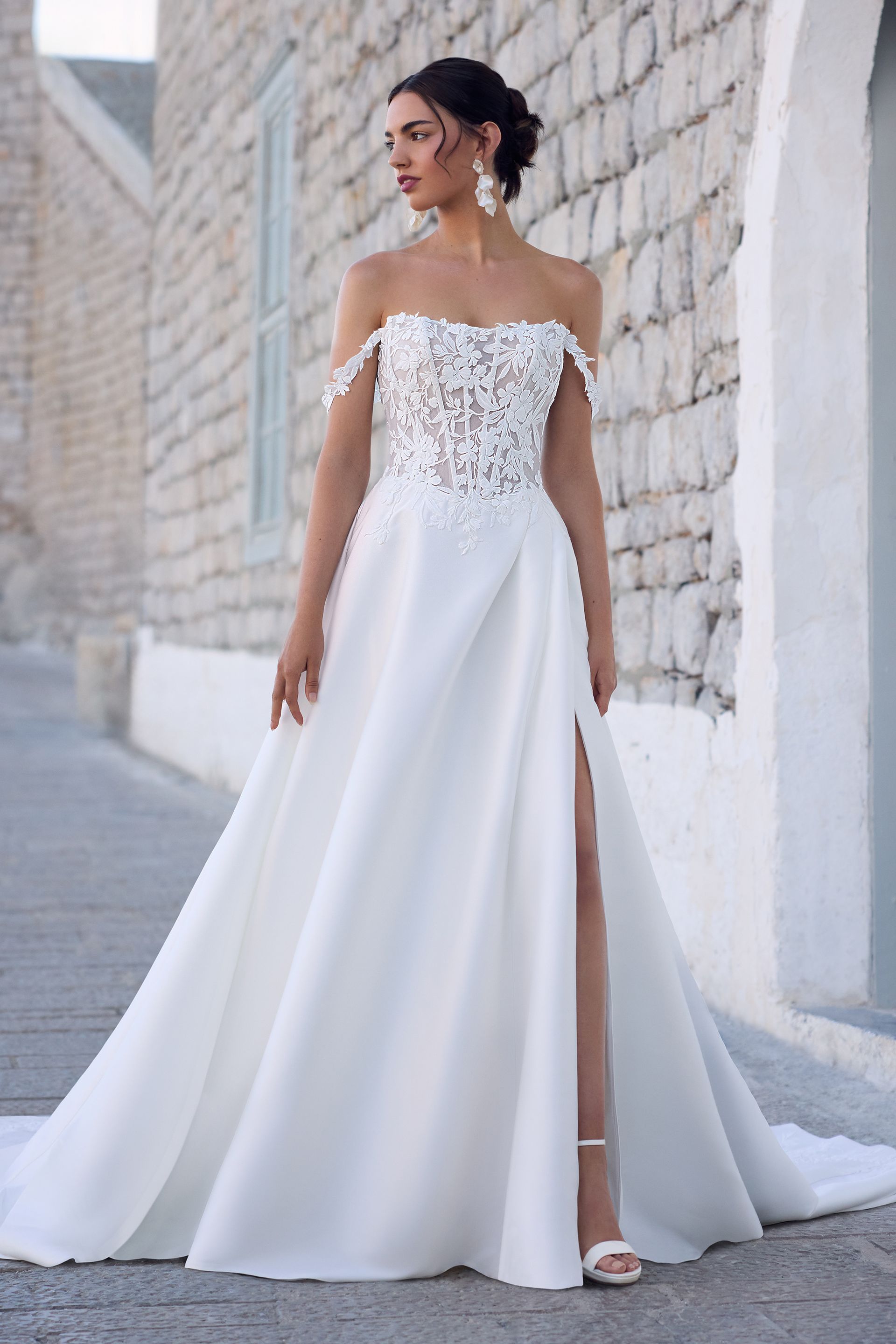 Woman in white off-the-shoulder wedding dress with high slit, standing outdoors.