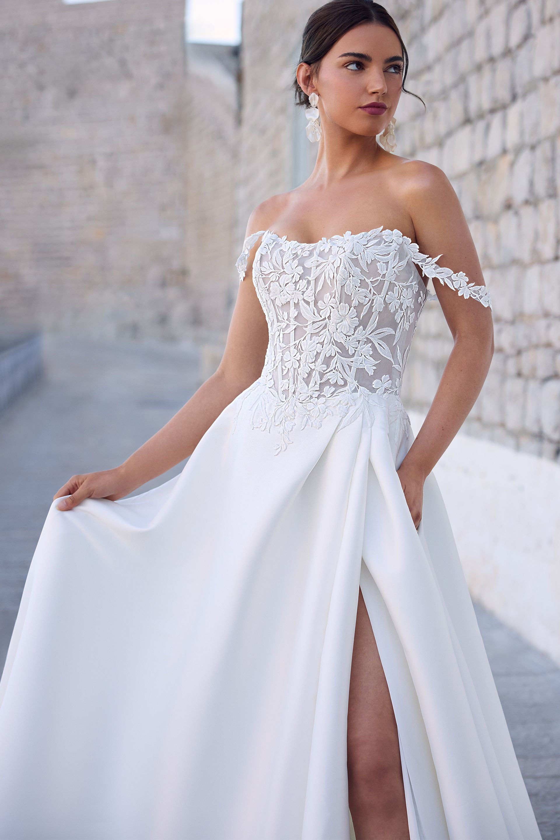 Woman in white off-the-shoulder wedding dress with high slit, standing outdoors.